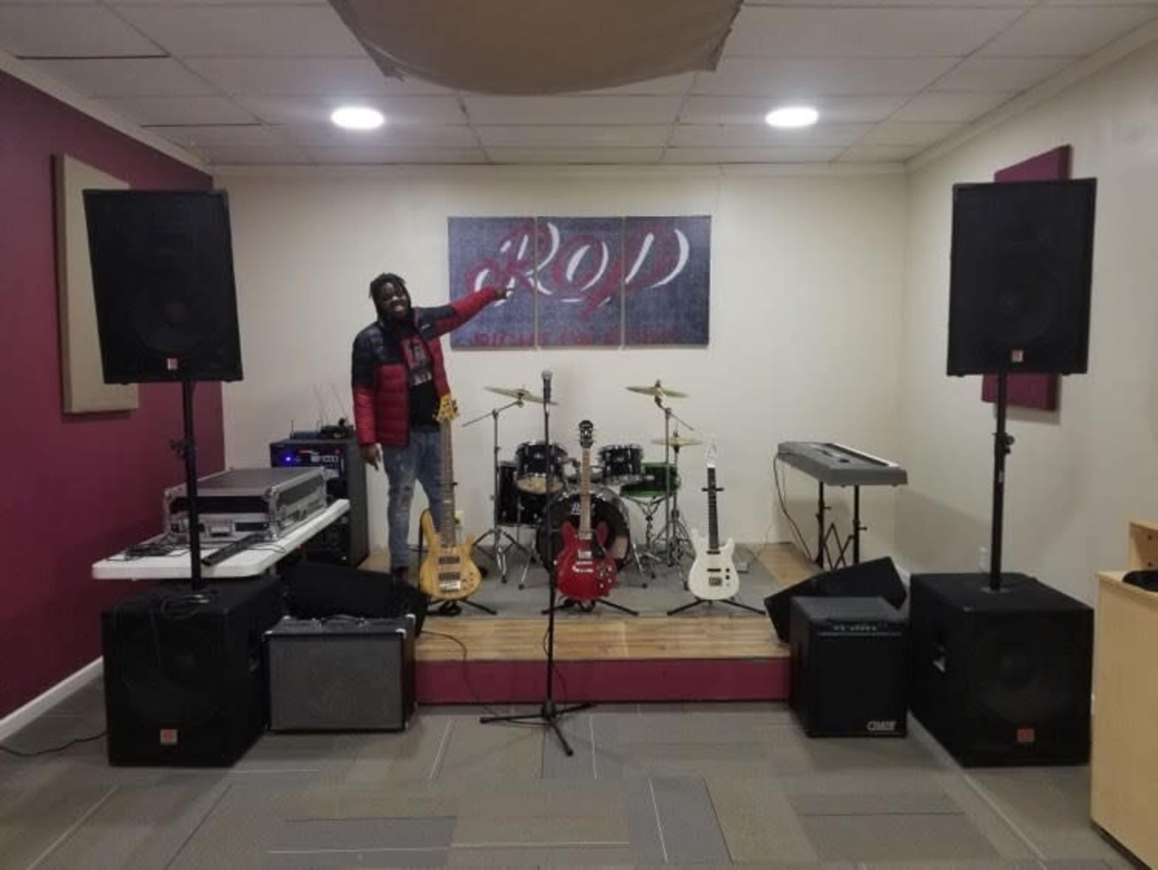 A man stands on a wooden stage in a music rehearsal space surrounded by musical instruments and audio equipment.