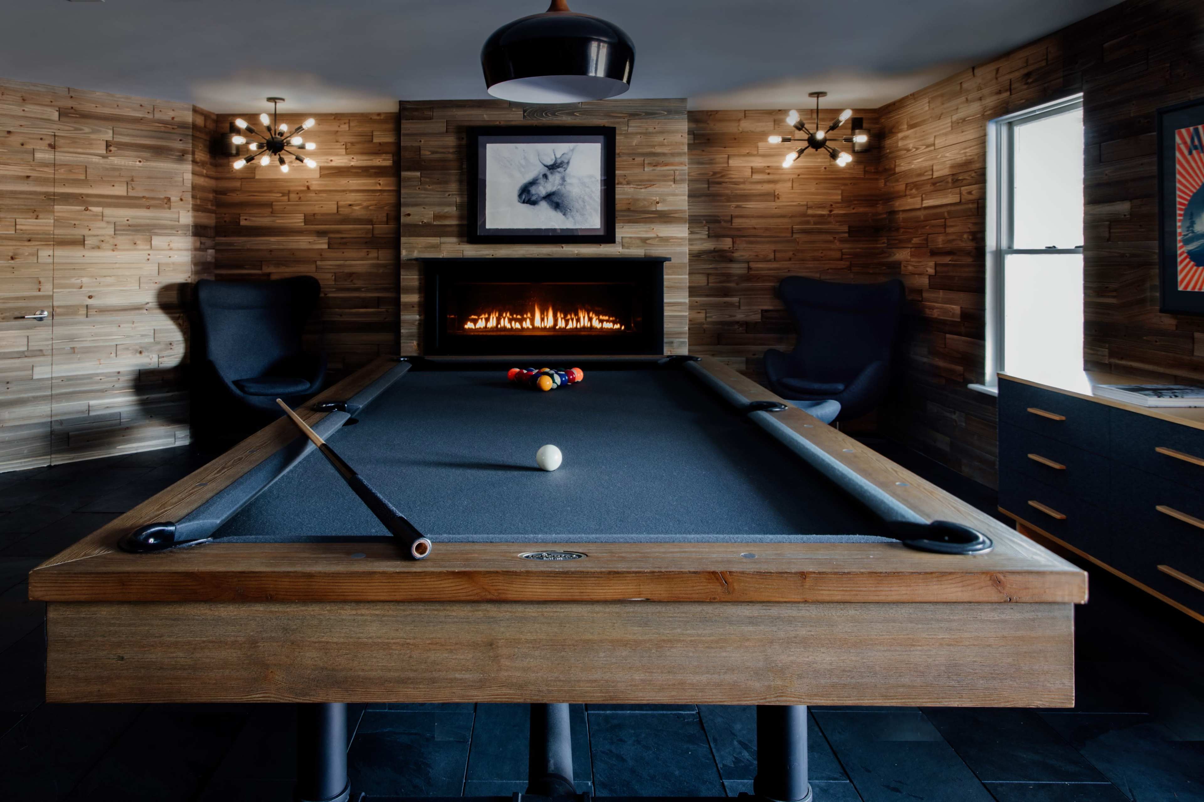 A billiard table with a white cue ball is positioned in a room featuring wooden walls, a fireplace, and modern lighting fixtures.