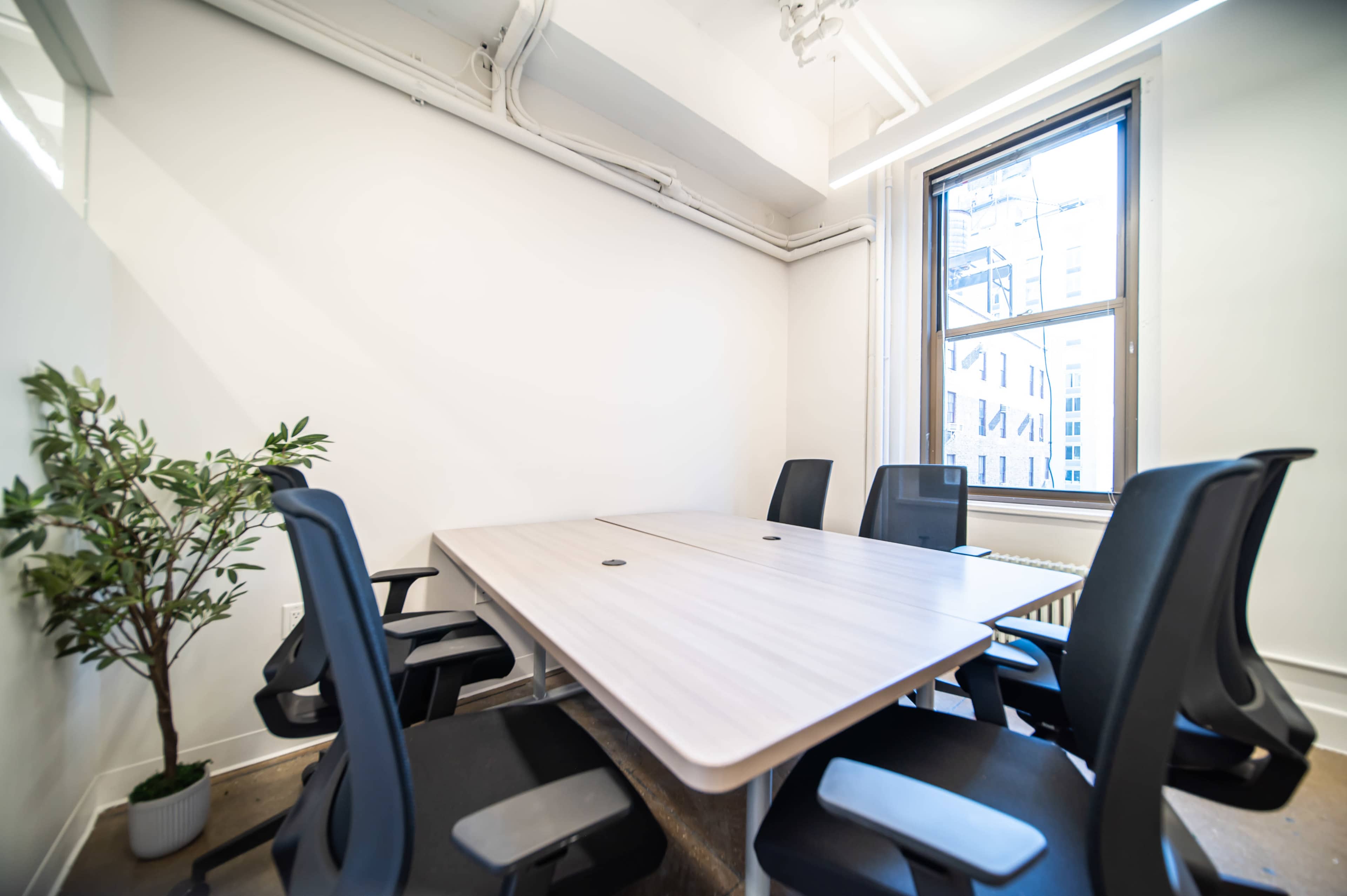 A small conference room features a light wooden table surrounded by six black ergonomic chairs and a potted plant near the window.