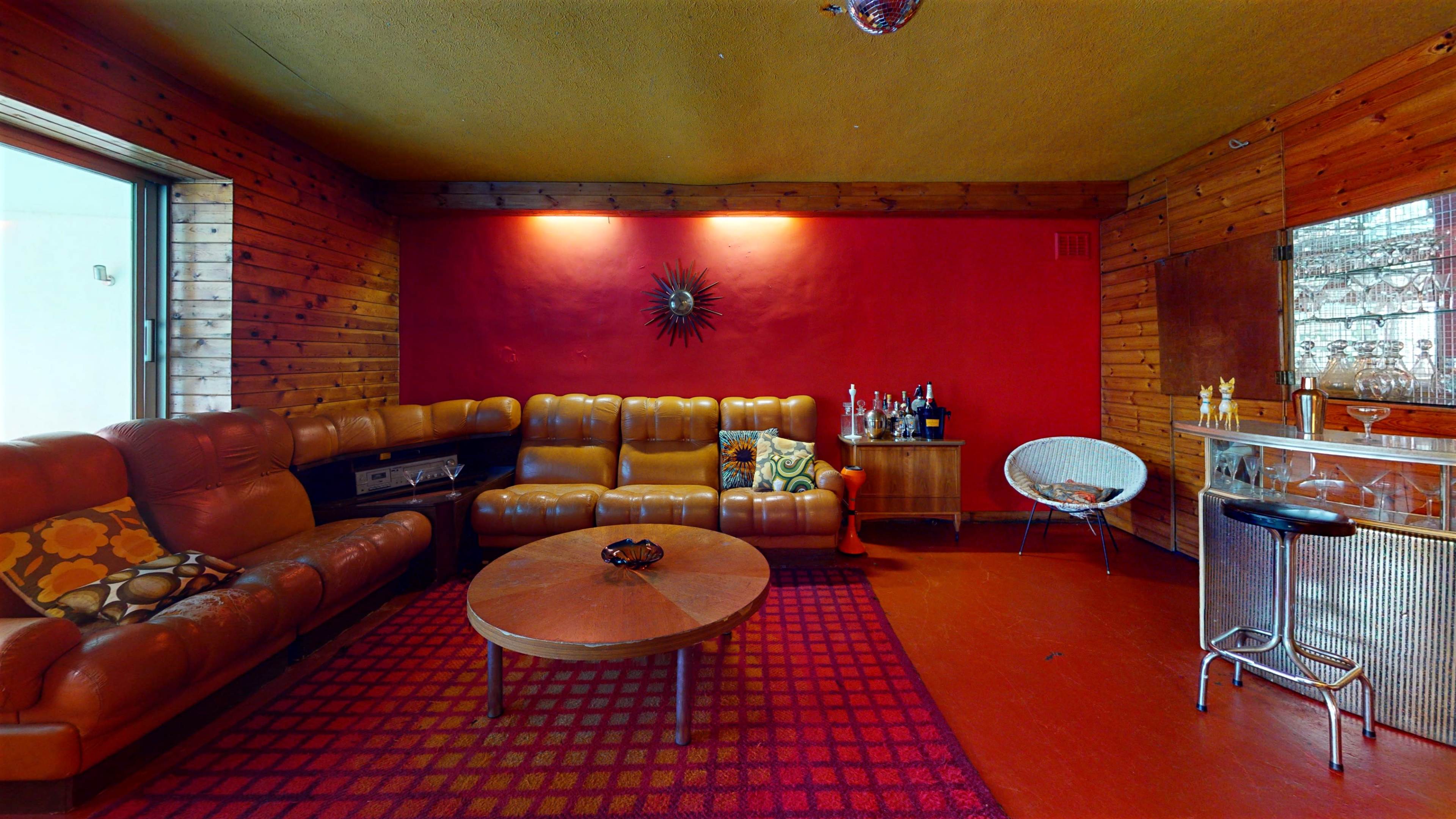 The image shows a cozy lounge with brown leather seating, a round wooden table, and a bar area against a wall adorned with wooden paneling and a bright red accent wall.