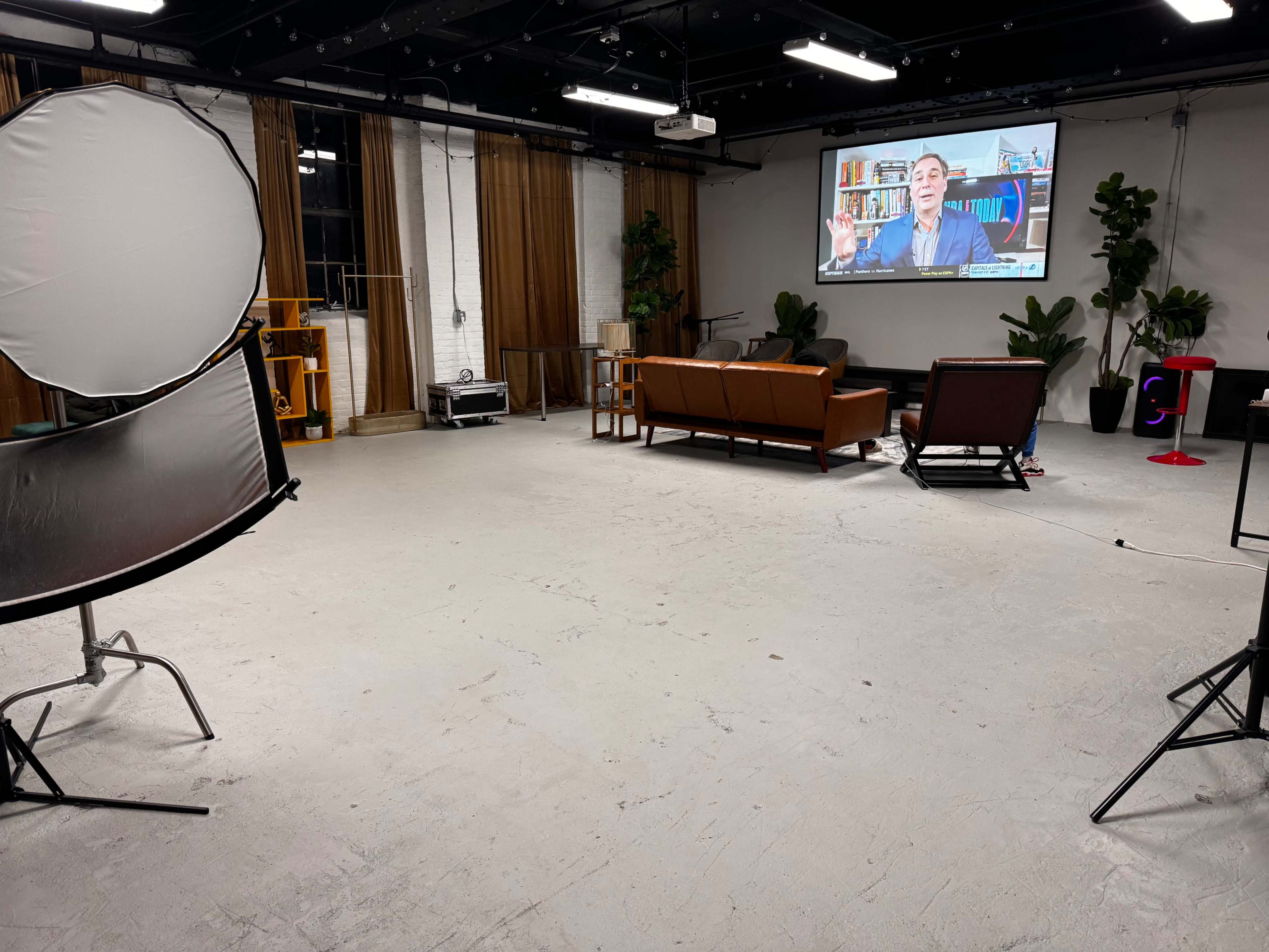 The image shows a spacious, well-lit studio with a large screen displaying a man speaking, two brown couches, and various plants arranged around the room.