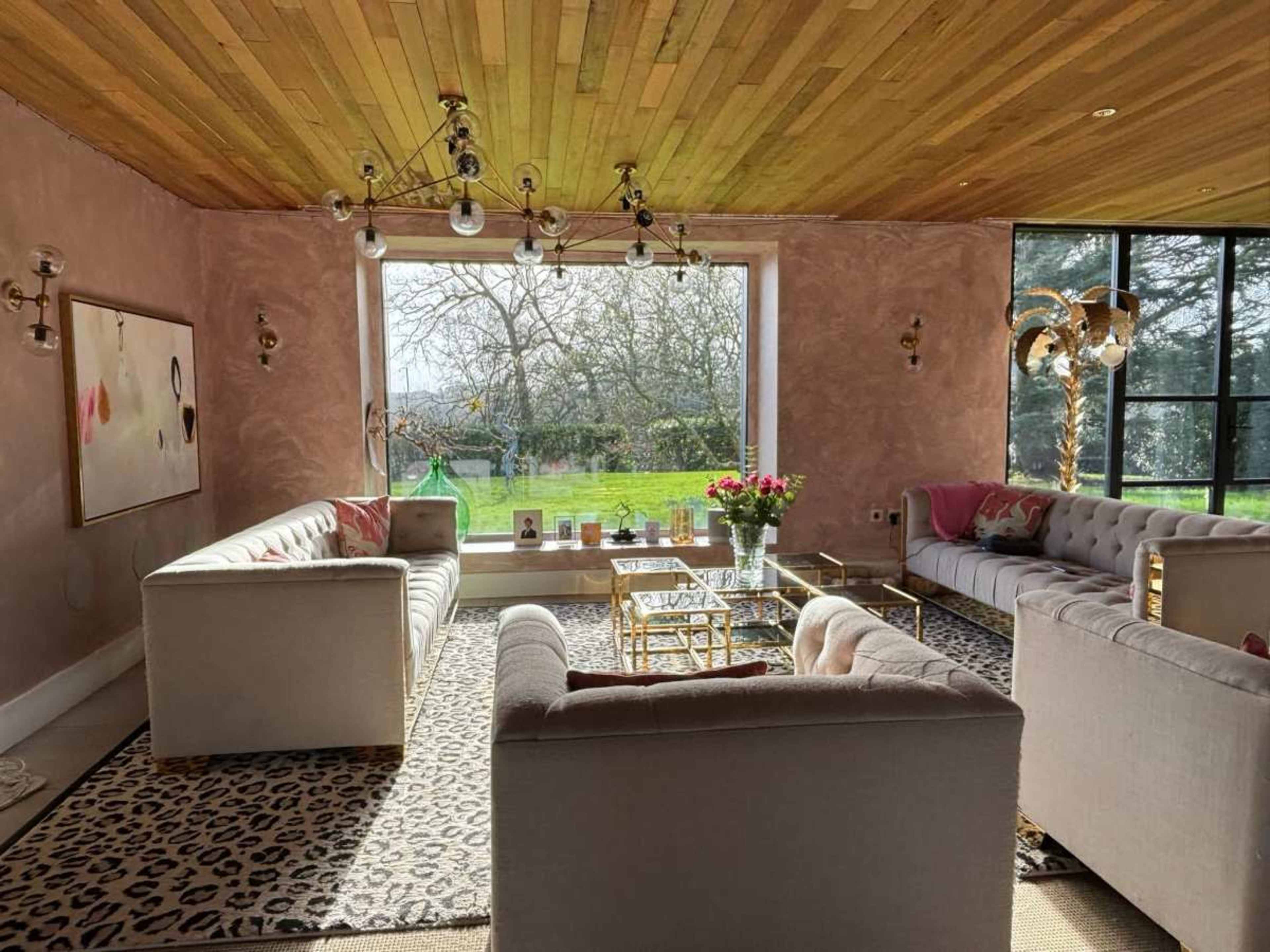 The image shows a spacious living room with large windows overlooking a garden, featuring two white sofas, a gold coffee table, and a leopard print rug.