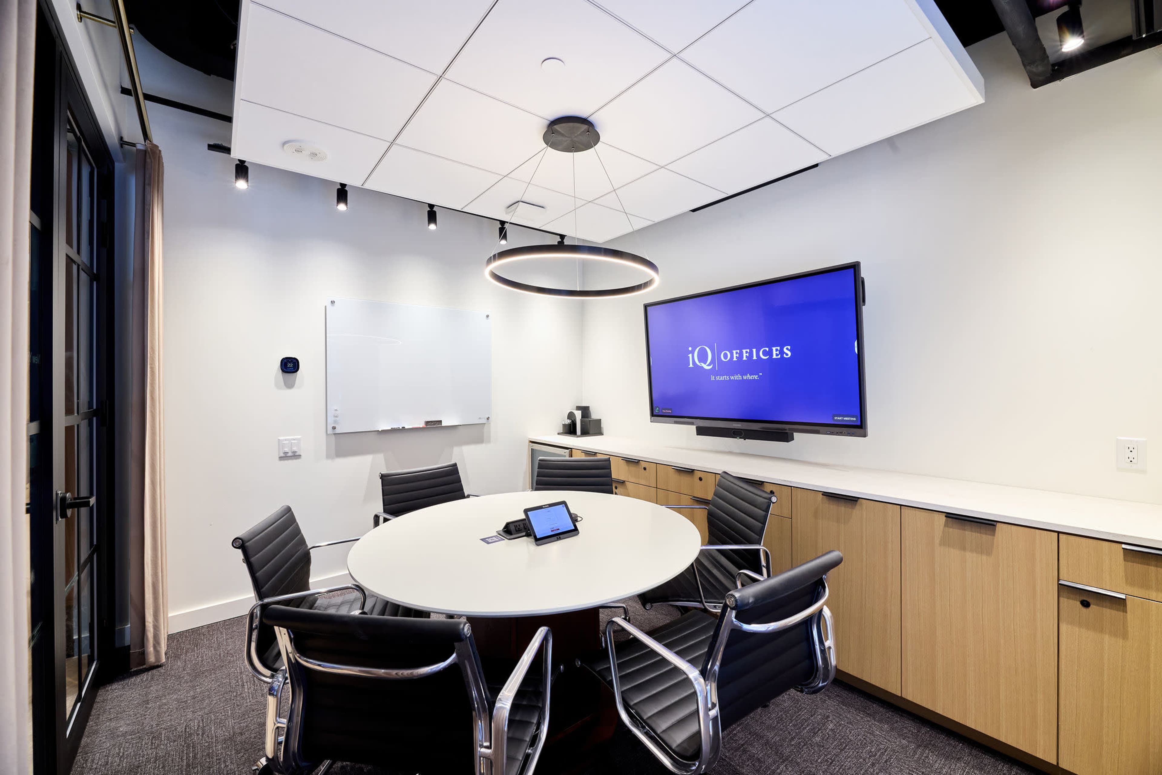 The image shows a modern conference room with a round table, black leather chairs, a wall-mounted screen, and a whiteboard.
