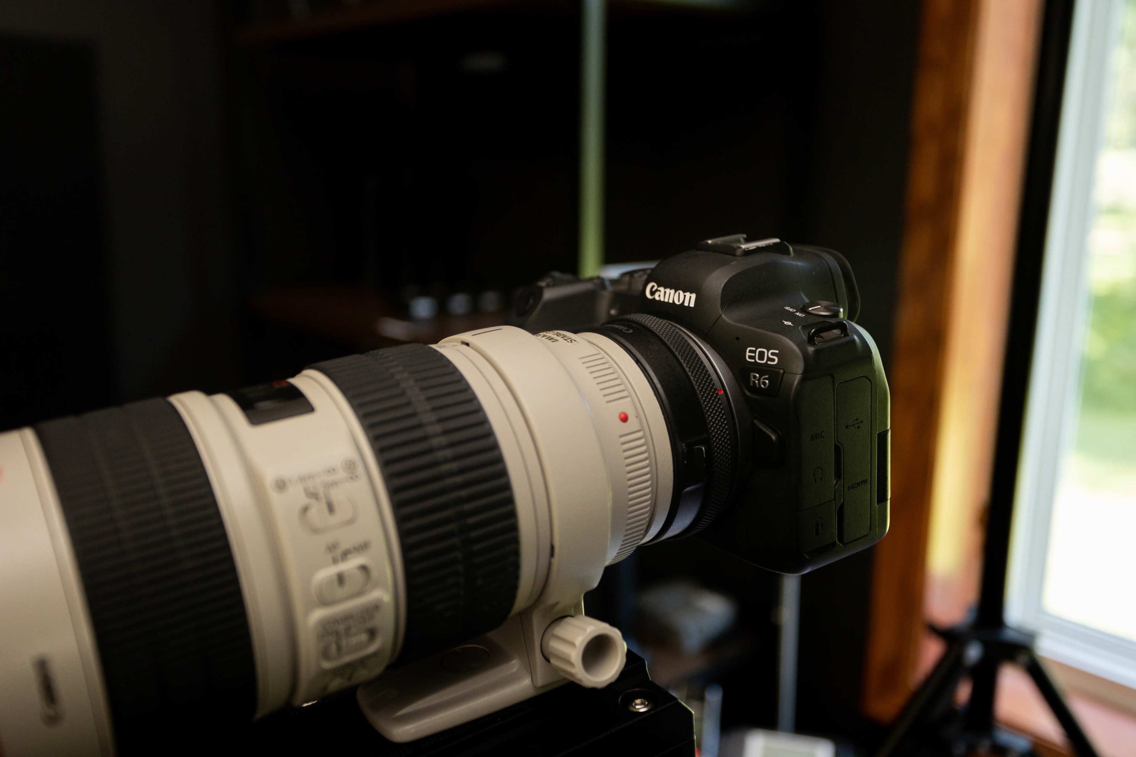 A Canon EOS R6 camera mounted with a large telephoto lens, positioned near a window with natural light.