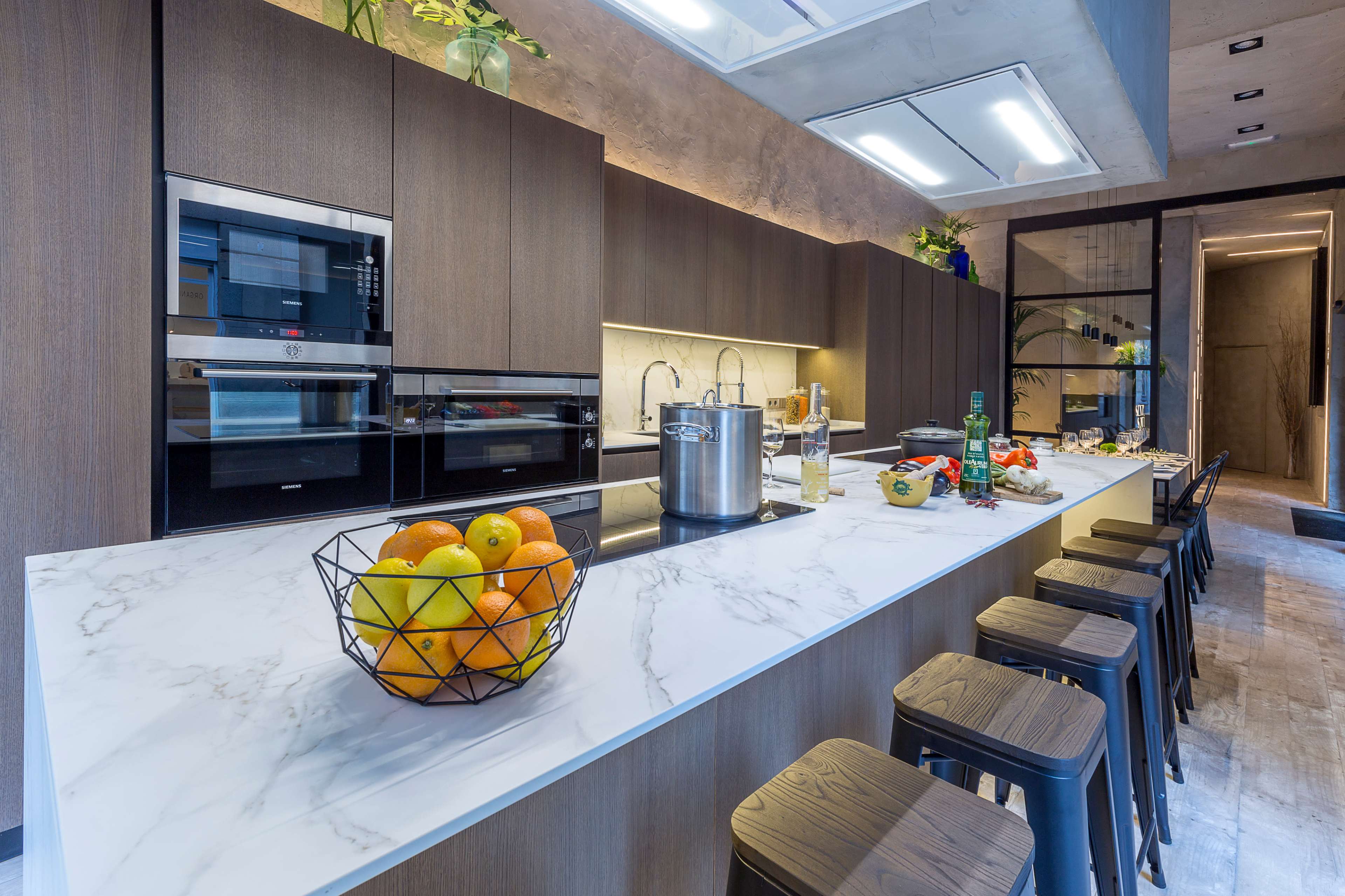A modern kitchen features a marble island with a fruit bowl, sleek wooden cabinetry, and stainless steel appliances.