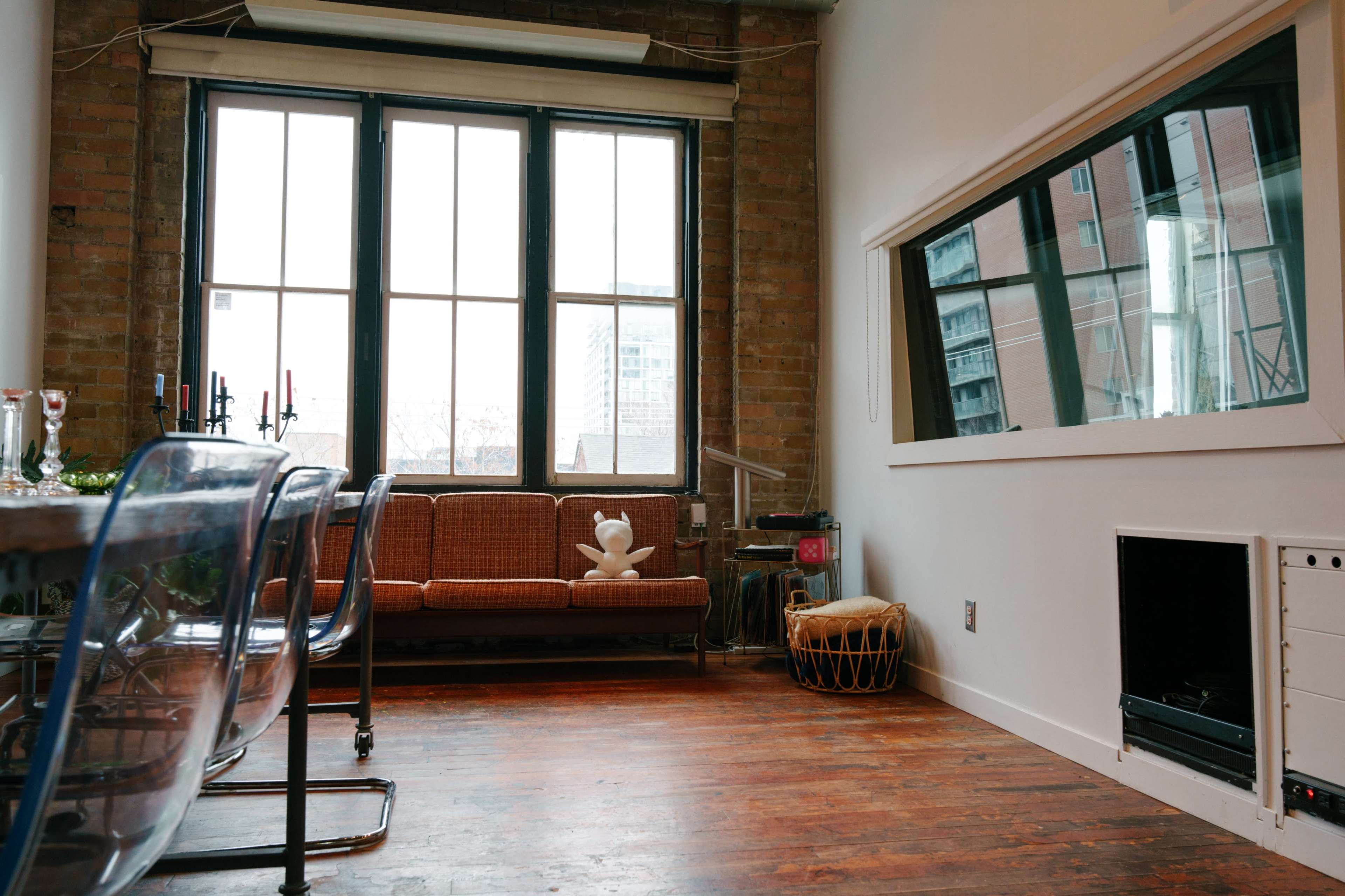 A spacious room with large windows, a vintage-style sofa, a dining area with transparent chairs, and a wall-mounted mirror.