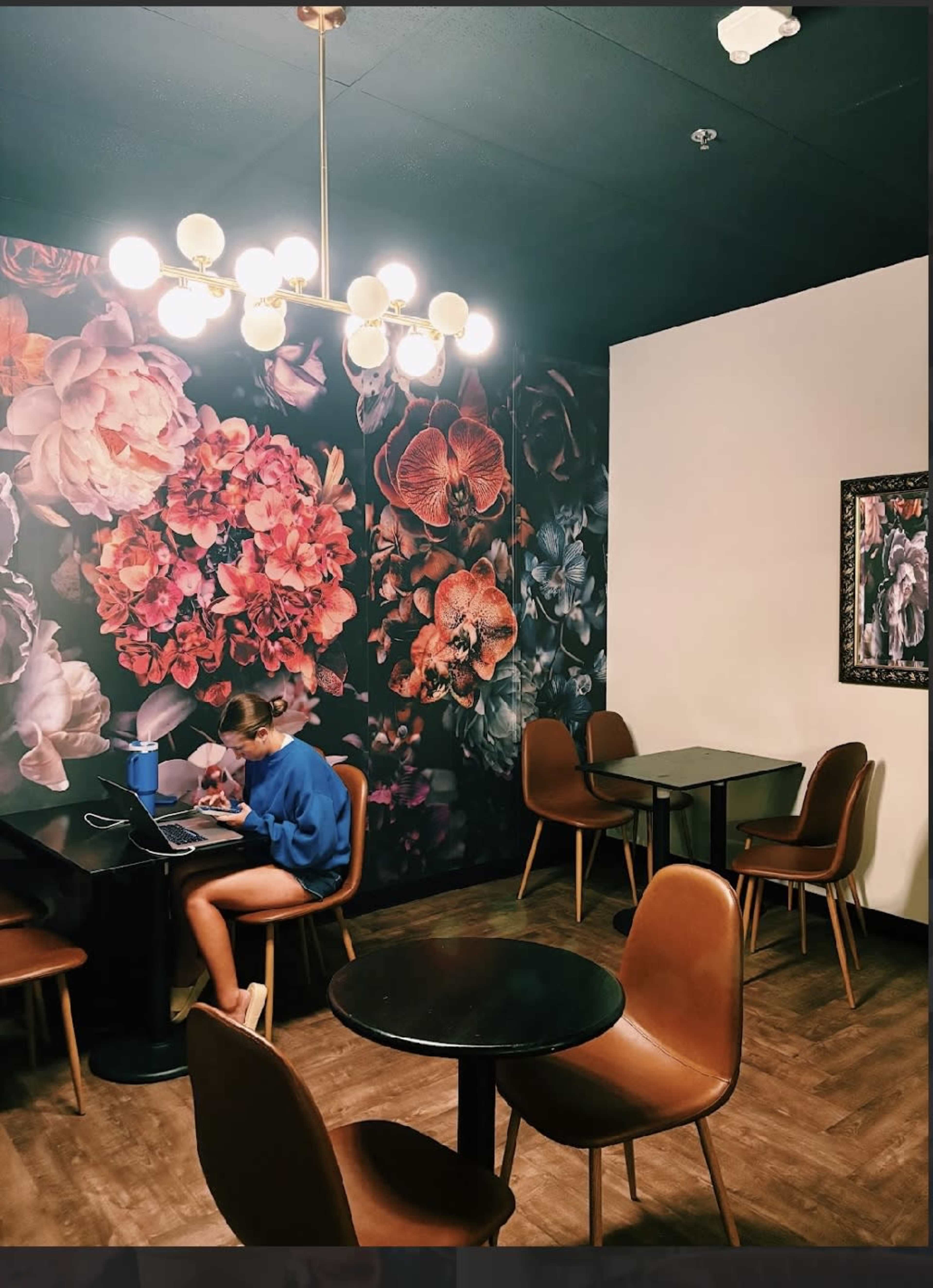A person is working on a laptop at a wooden table in a cafe with floral wallpaper and minimalist furniture.