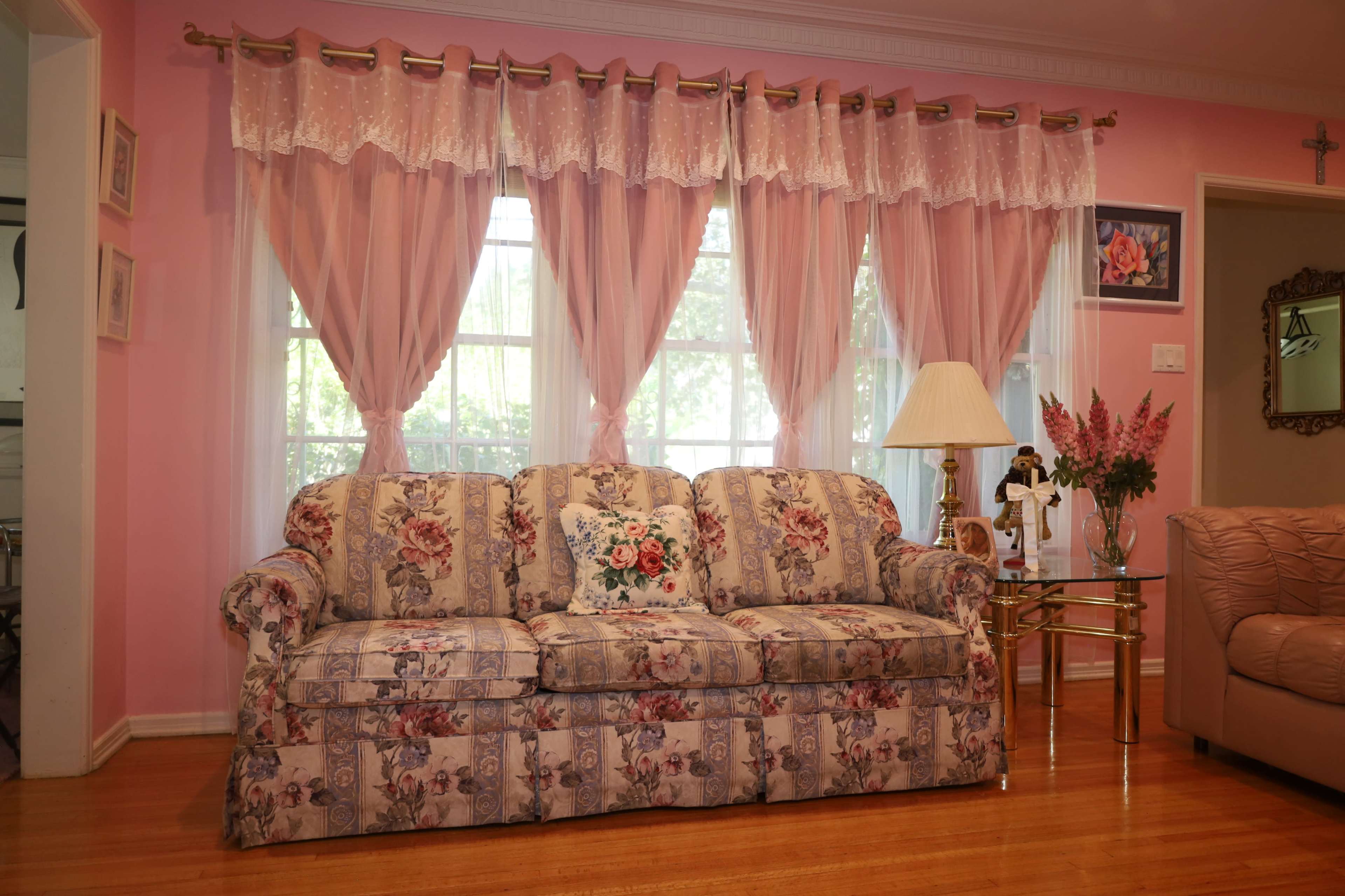 The image shows a living room with a floral-patterned sofa, light pink curtains, a lamp, and a decorative table.