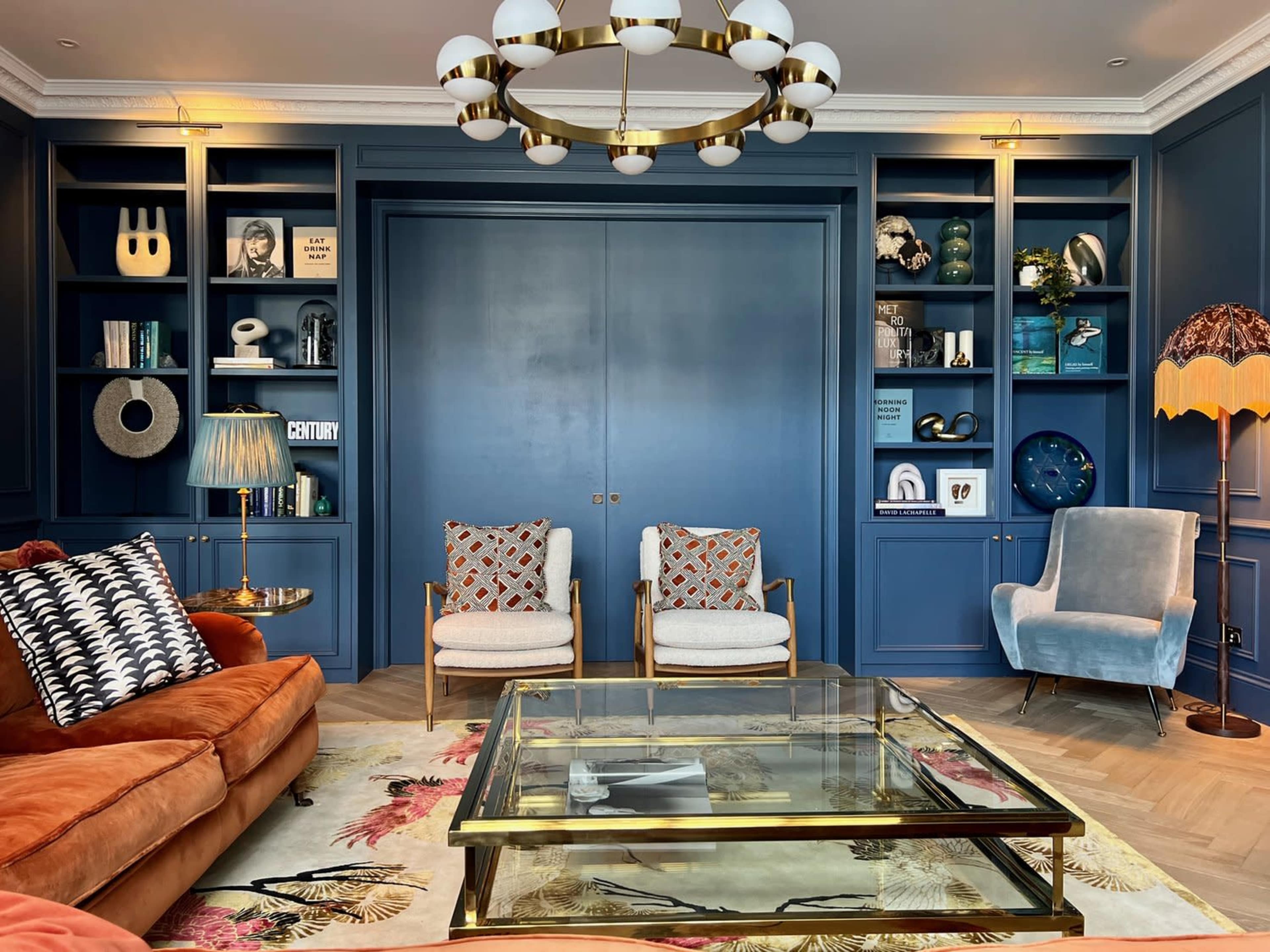 A stylish living room with a navy blue feature wall, a glass coffee table, two armchairs, a large orange sofa, and shelves filled with decorative items and books.