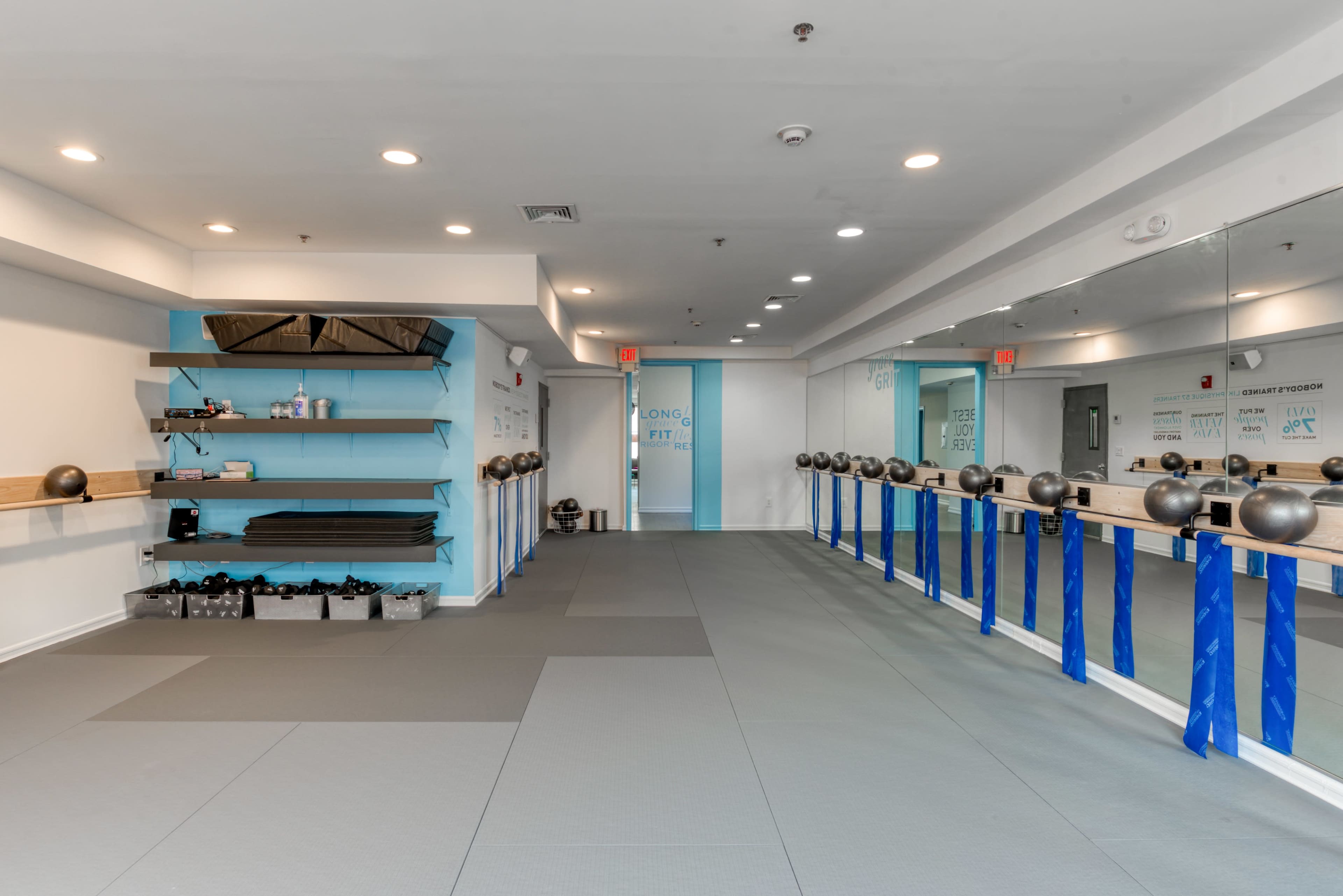 The image shows a spacious, well-lit fitness studio with gray flooring, wall-mounted shelves containing exercise equipment, and a row of kettlebells lined up along a mirrored wall.