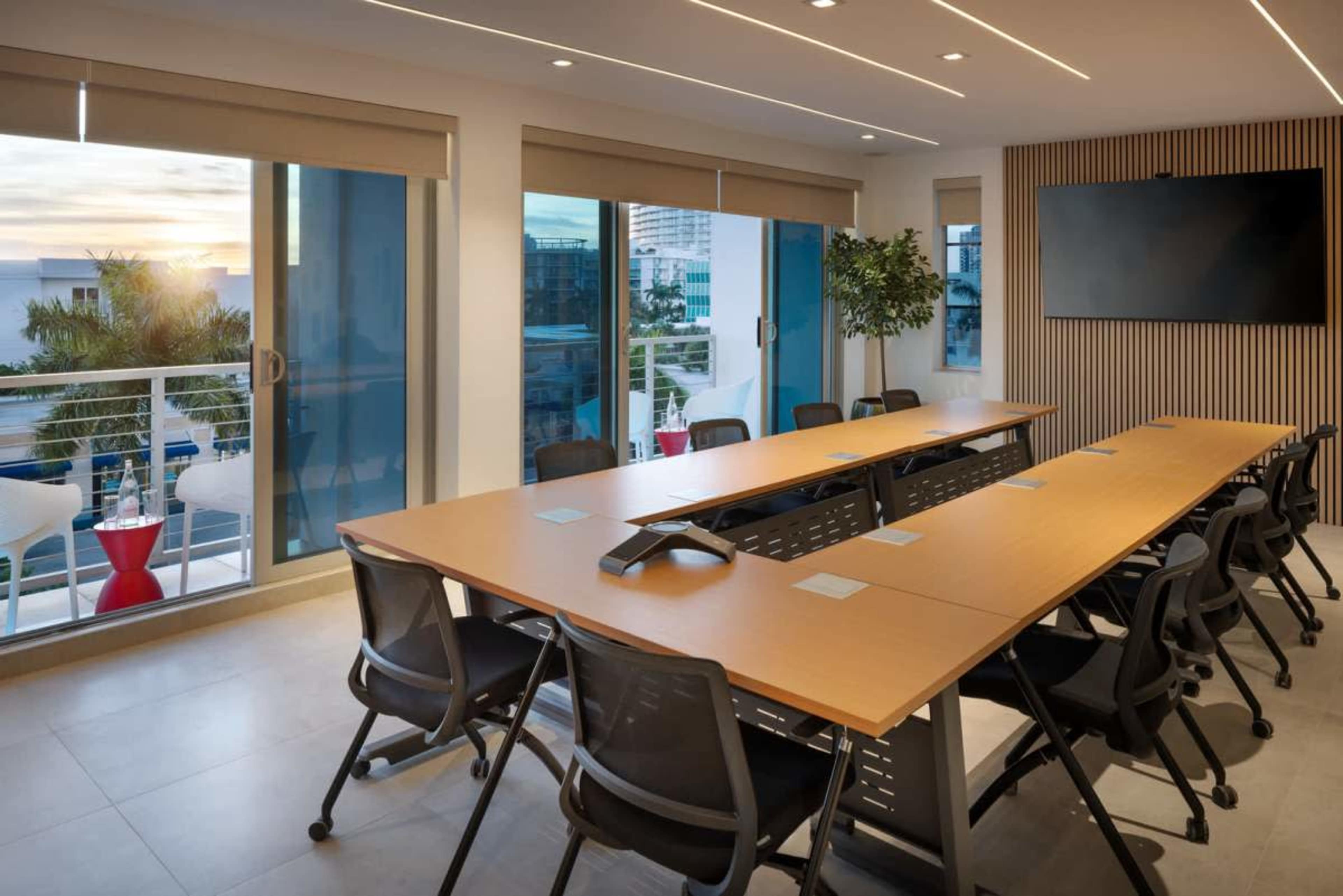 A modern conference room features a long wooden table with ergonomic chairs, large windows that allow natural light to illuminate the space, and a mounted screen on the wall.