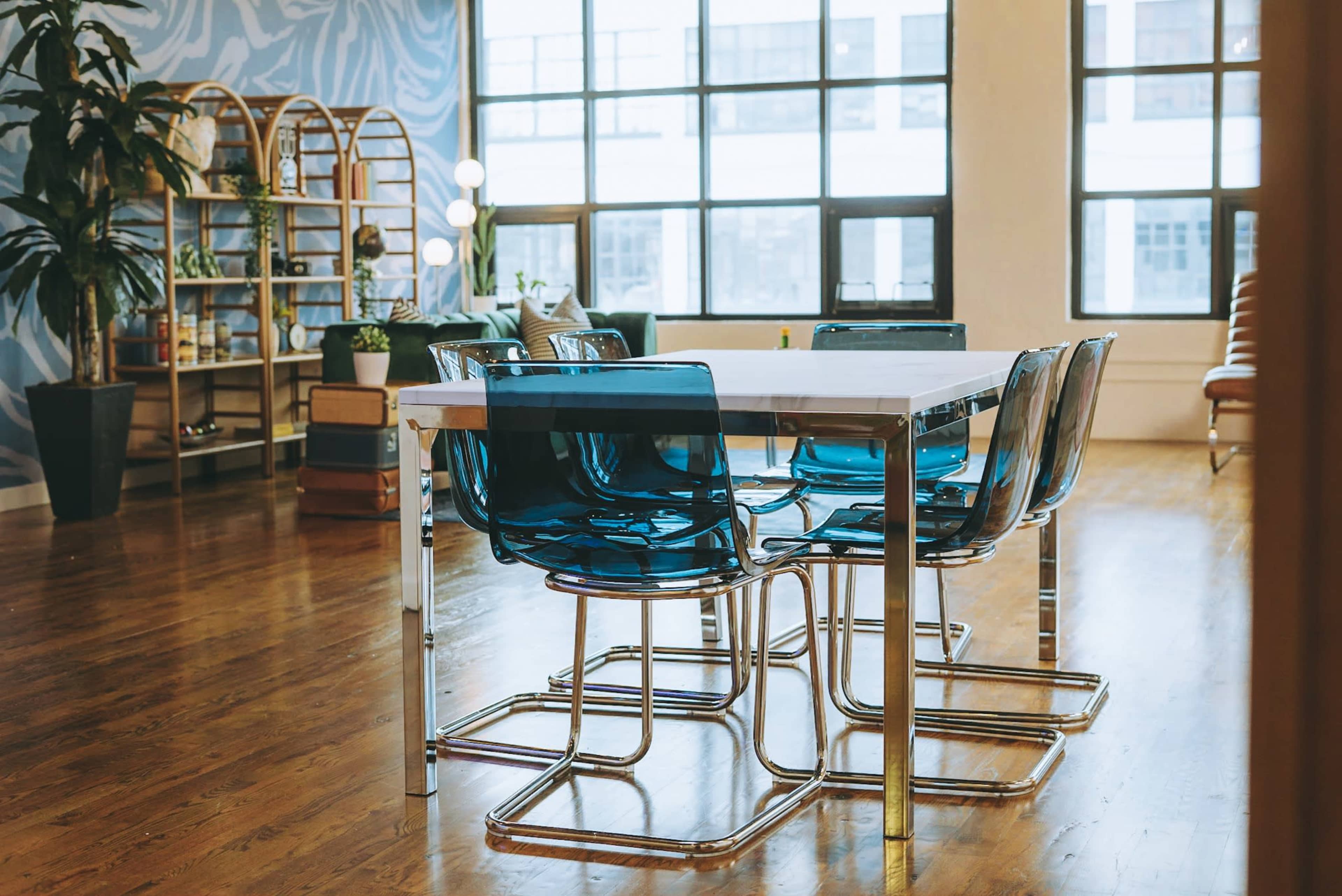 A modern dining area features a white table surrounded by six transparent blue chairs, with large windows allowing natural light to fill the space.