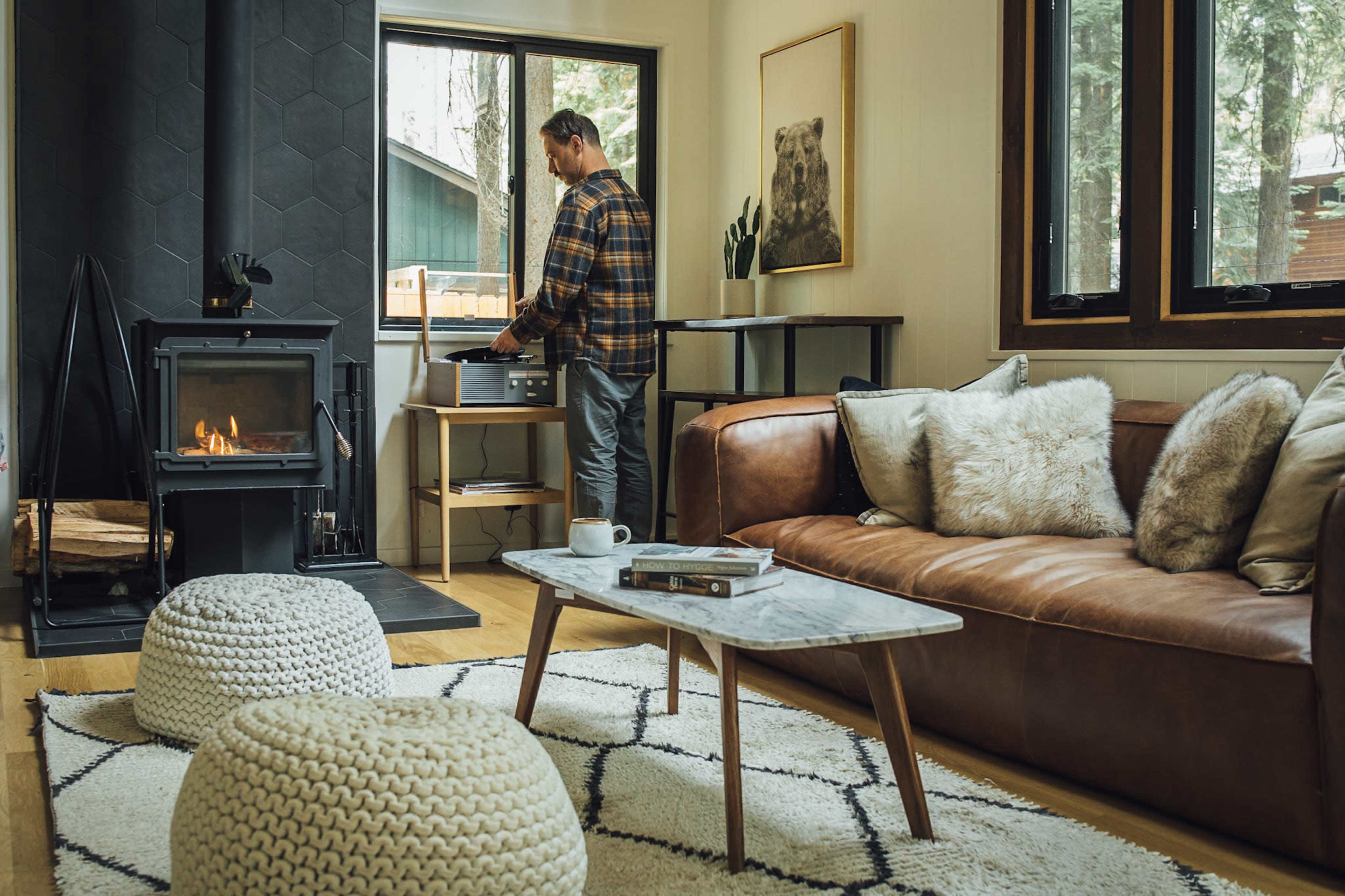 A man stands by a record player next to a large window in a cozy living room with a fireplace and a leather sofa.