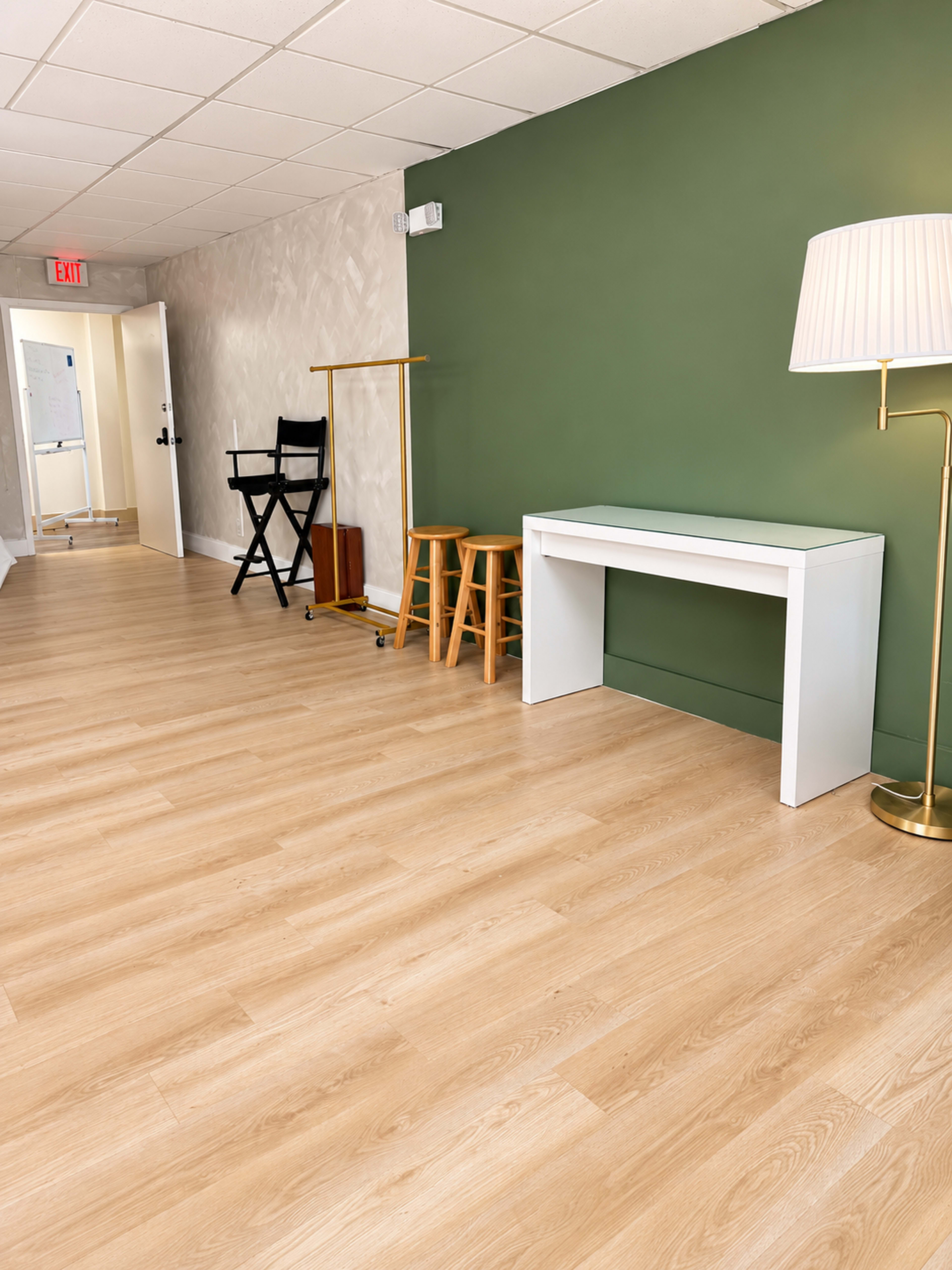 The image shows a hallway with light wood flooring, a green accent wall, a white console table, and a lamp, along with a gold-hued stool and a clothing rack in the background.