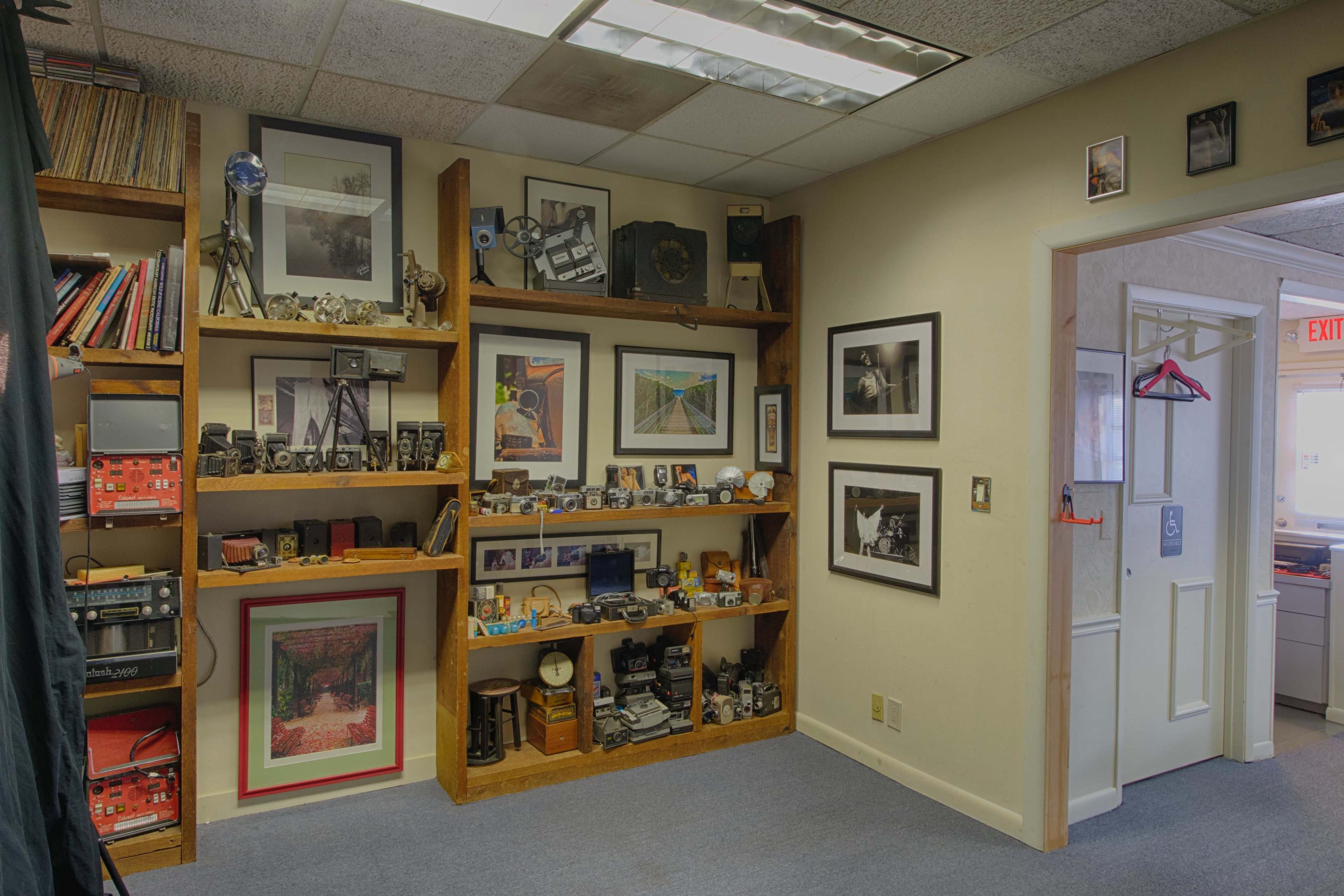 The image shows a corner of an office with wooden shelves displaying various photographs, cameras, and decorative items, adjacent to an open doorway leading to another room.