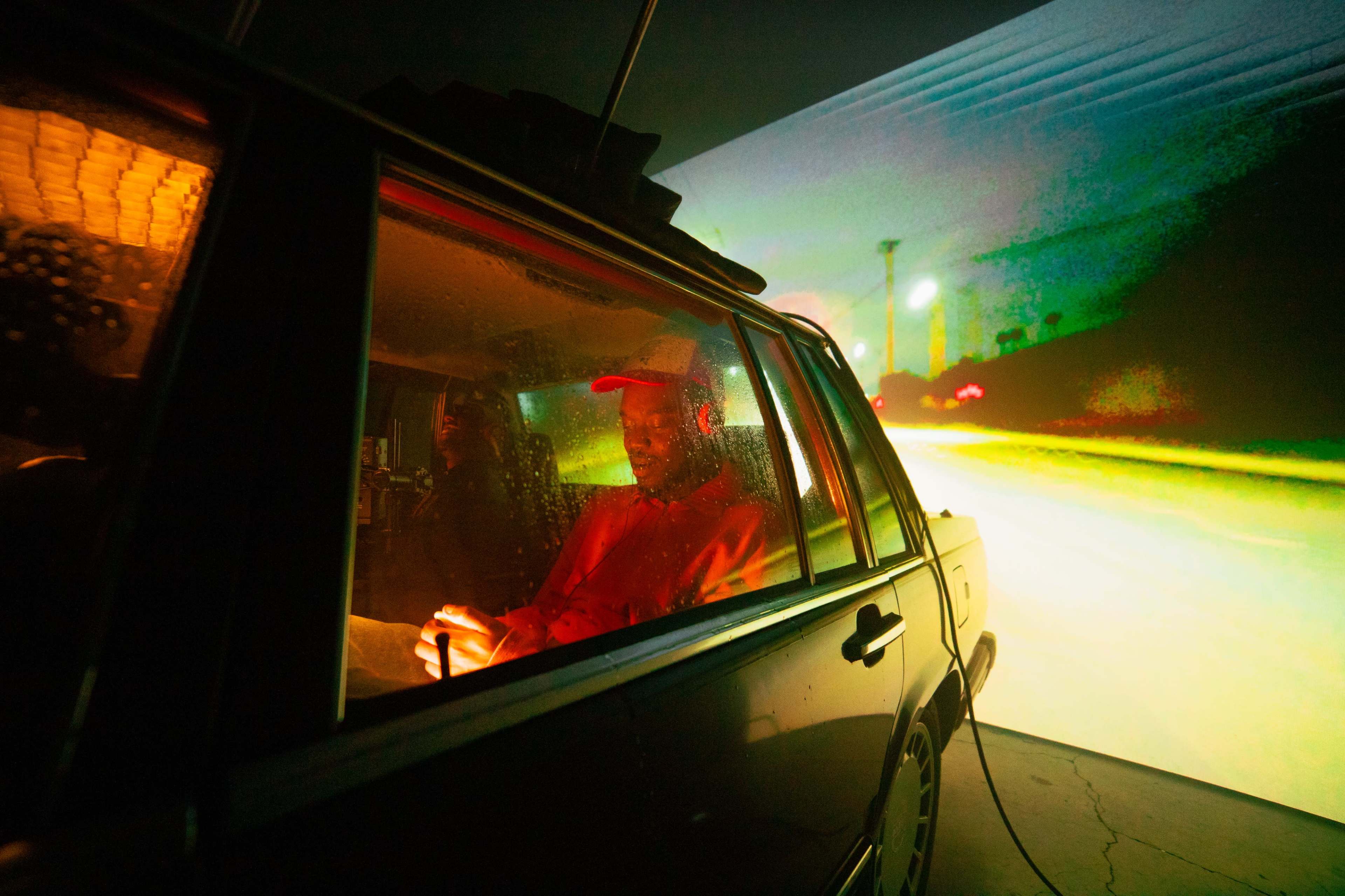 A person sits inside a car with colorful lighting, while a vibrant projection of a night scene illuminates the background.