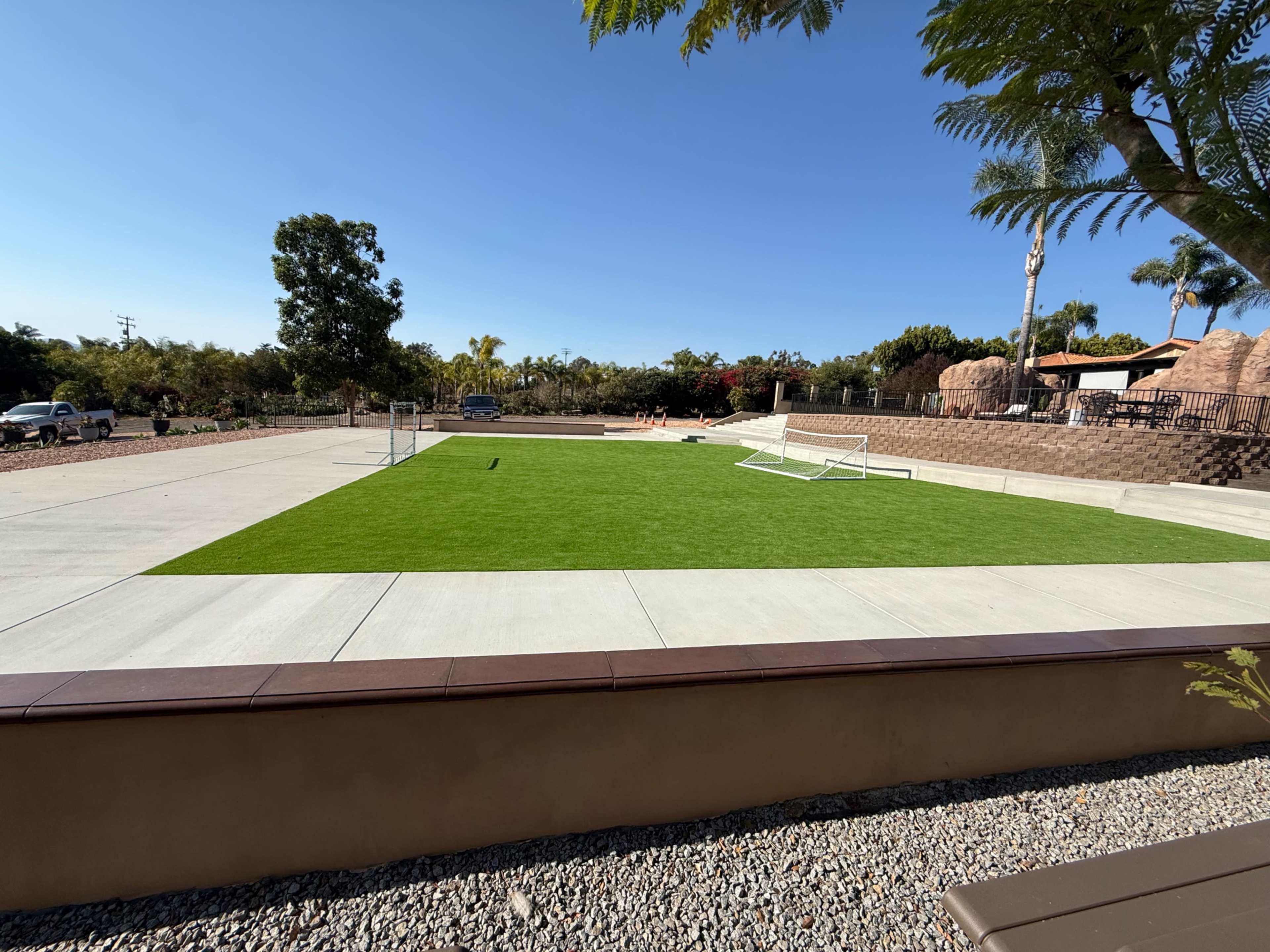The image shows a spacious, green artificial turf area in a park setting, surrounded by concrete pathways and landscaping.