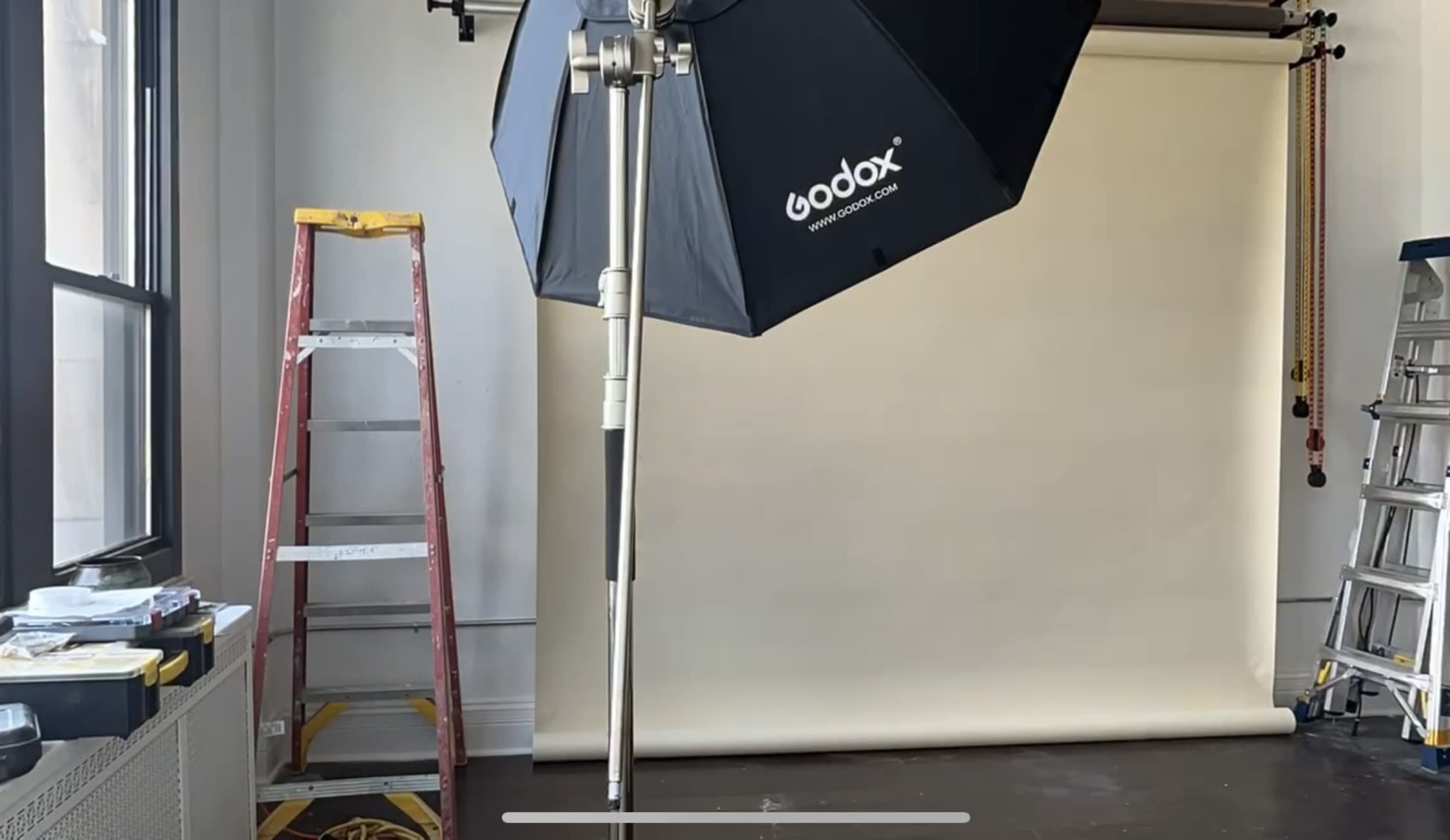 A Godox light umbrella is positioned next to a wooden ladder in a photography studio with a neutral backdrop.