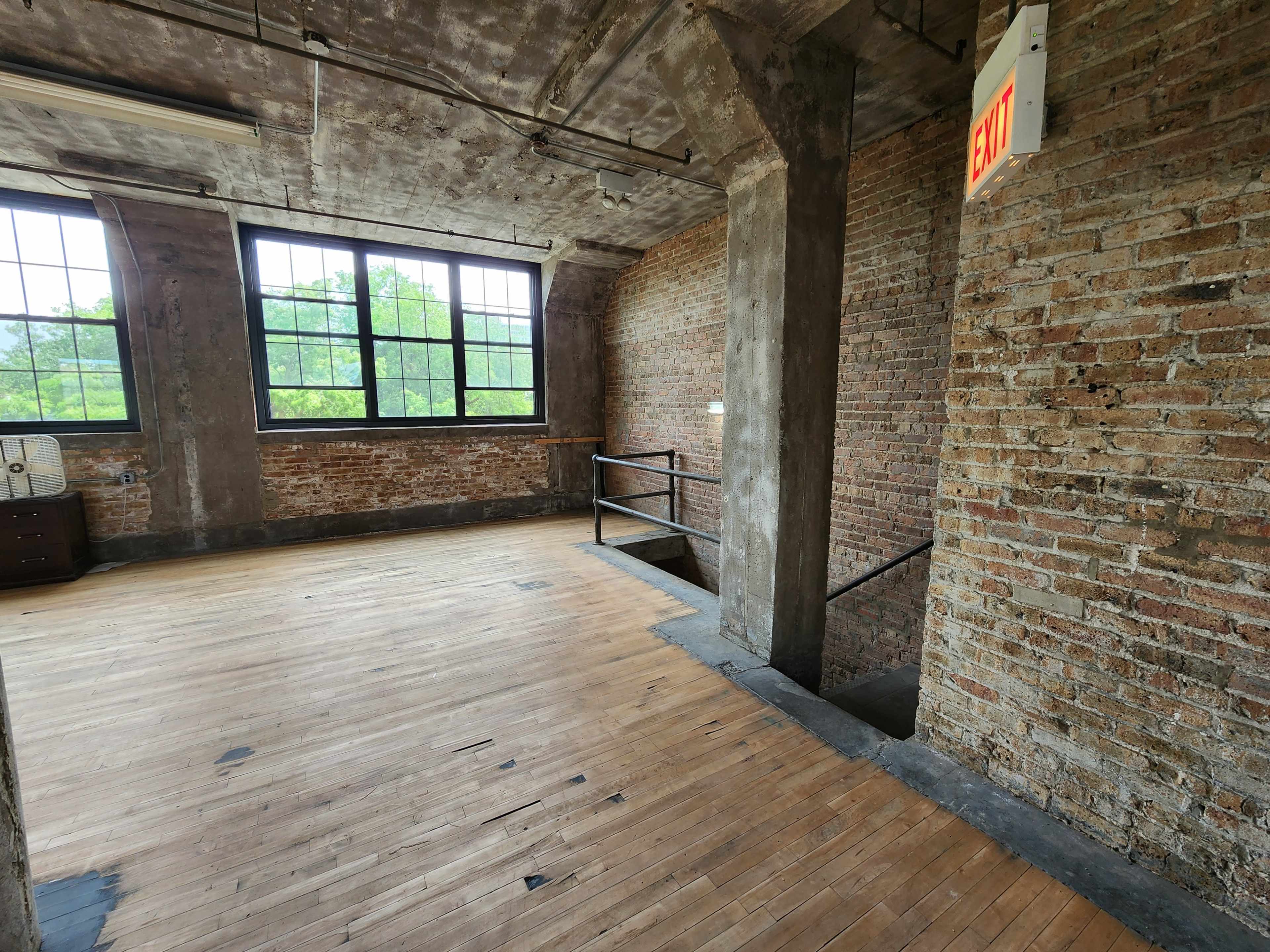 An empty room with exposed brick walls, wooden floors, large windows, and an exit sign near a staircase.