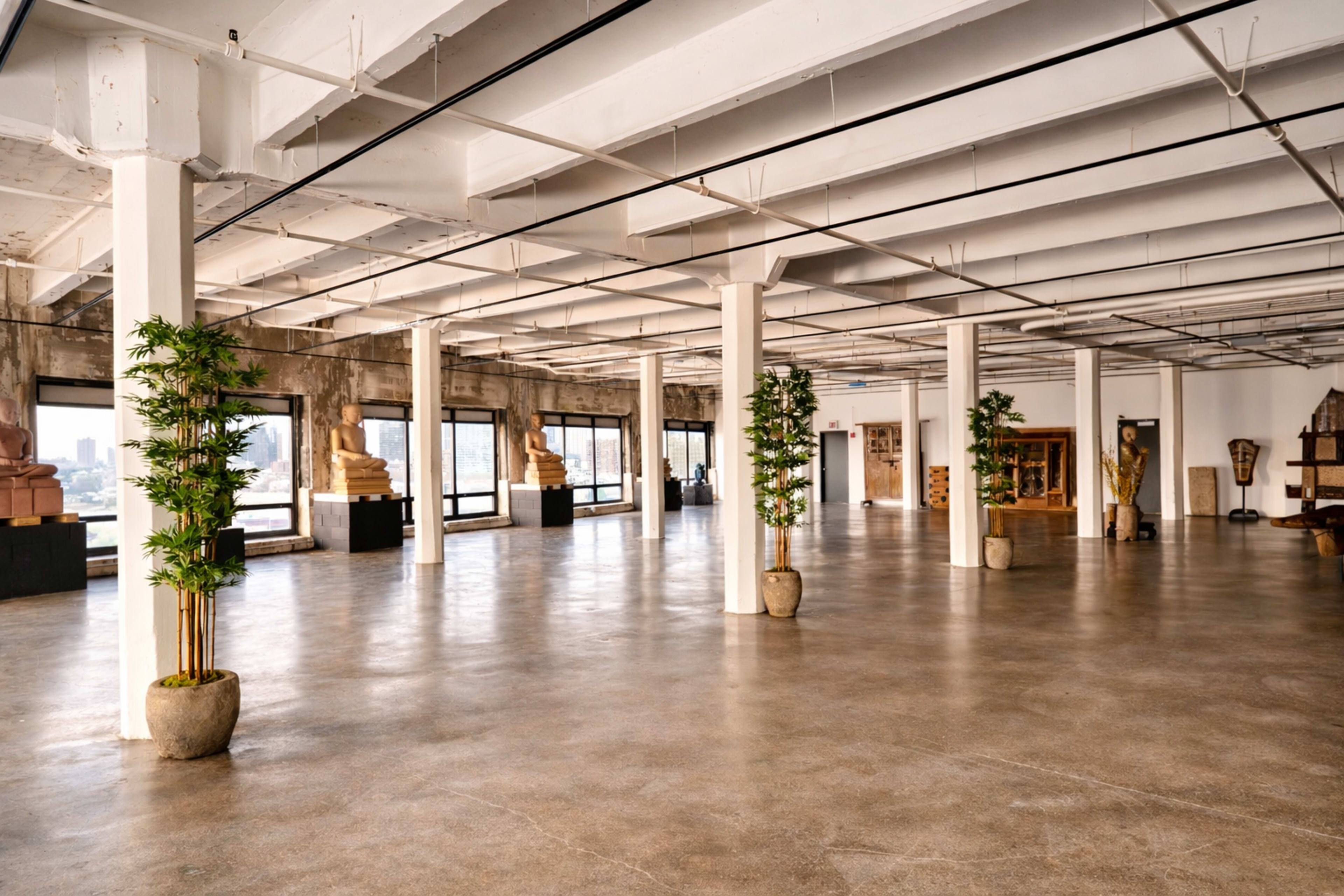 The image shows a spacious gallery with polished concrete floors, white columns, and large windows, featuring several sculptures displayed on pedestals.