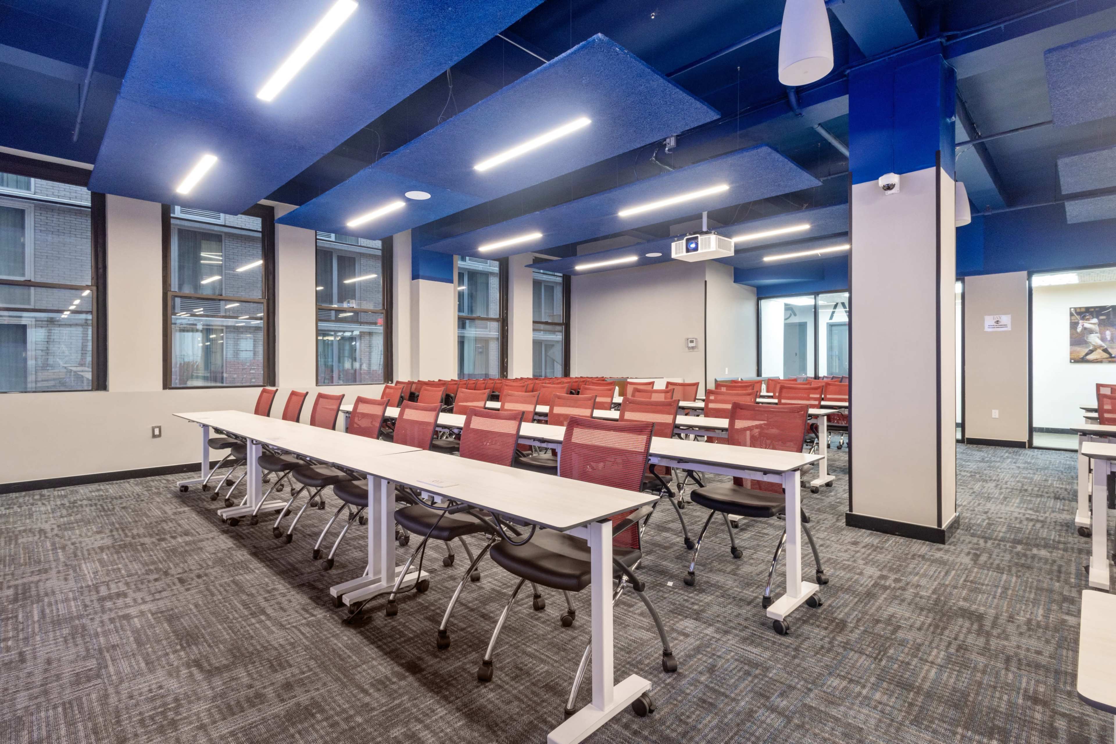 A modern classroom features rows of red and black chairs with white tables, illuminated by overhead lights and equipped with a ceiling-mounted projector.