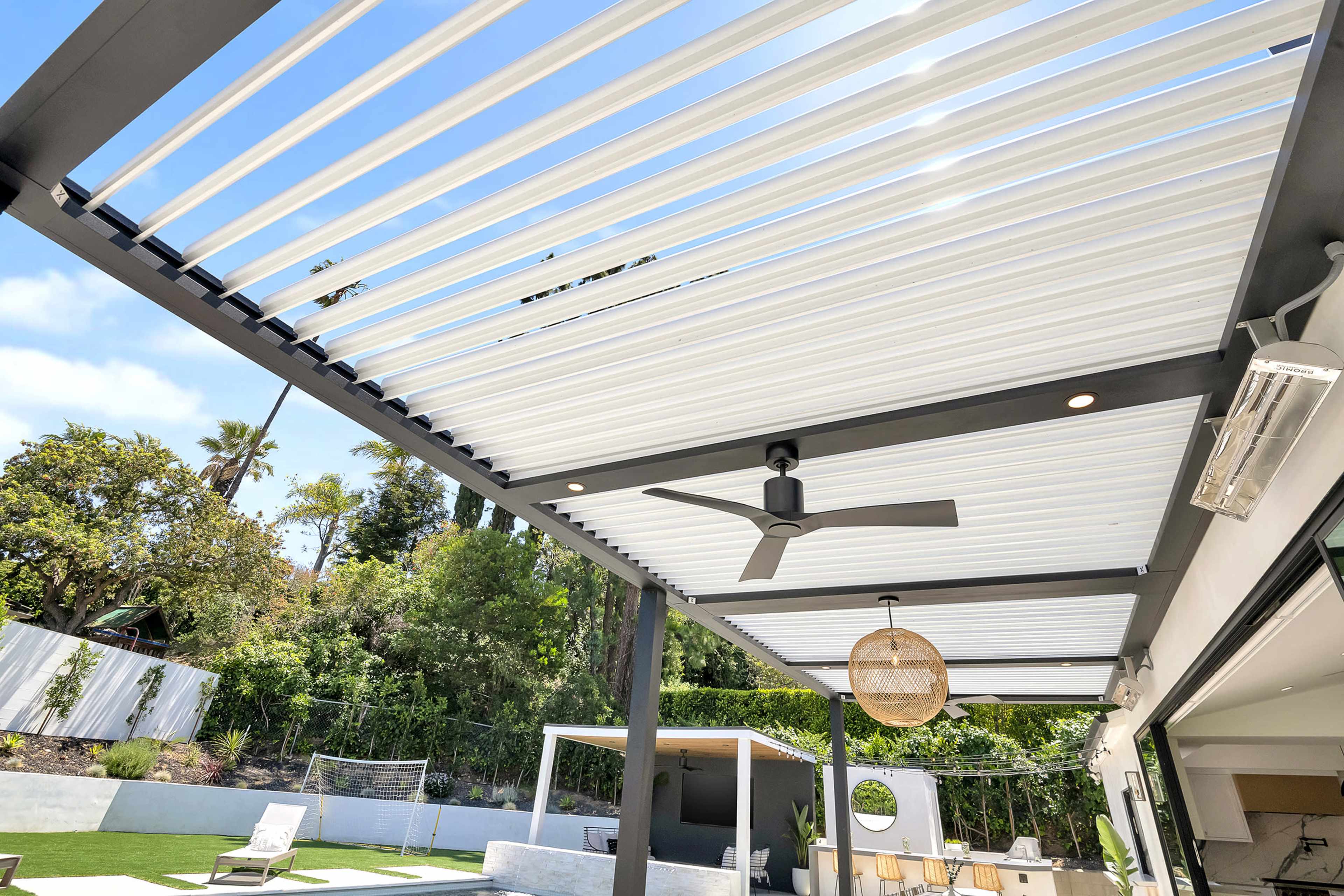 A patio with a slatted roof, outdoor seating, and greenery in the background.
