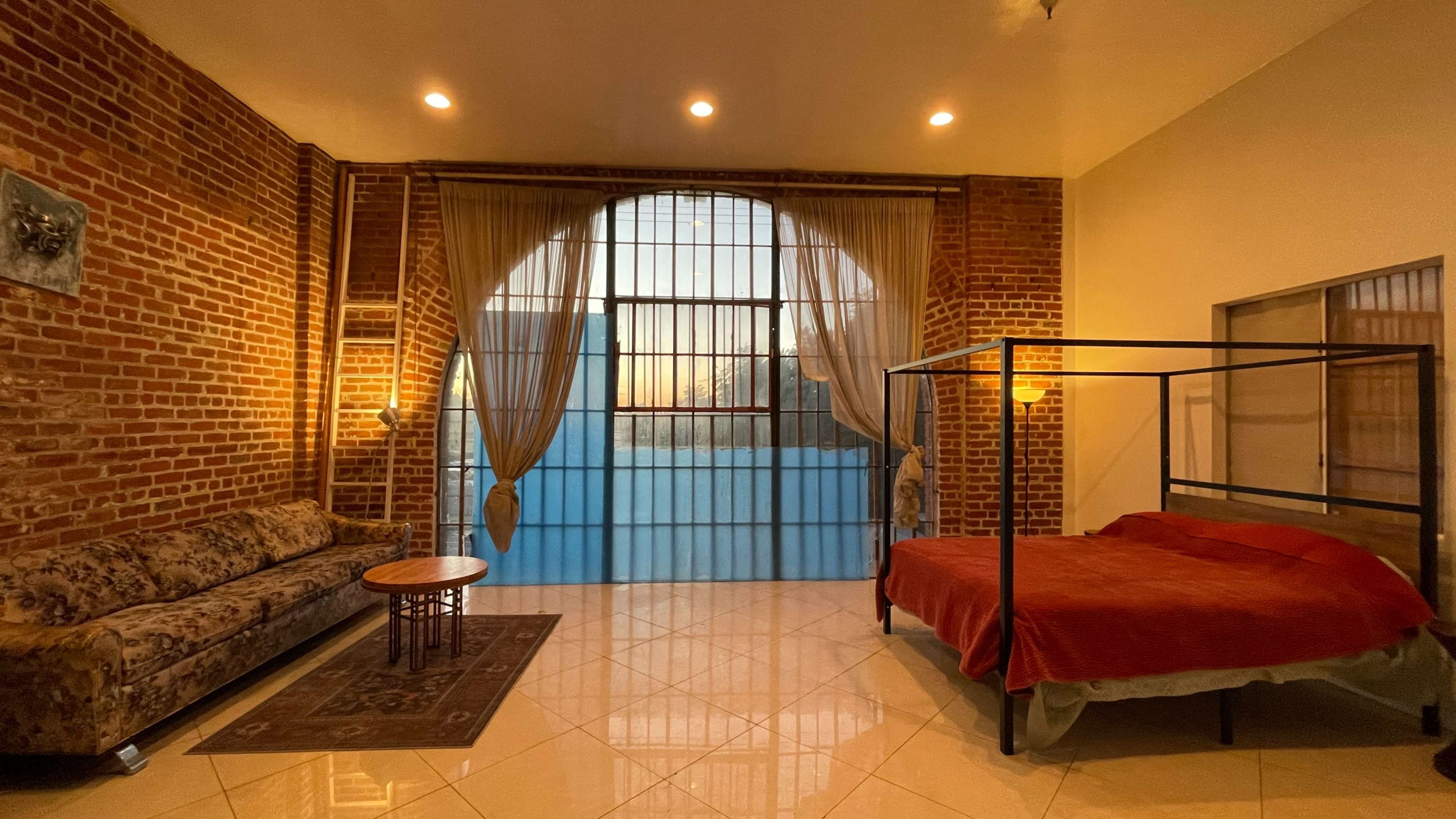 A spacious bedroom features a brick wall, a large window with curtains, a four-poster bed, and a vintage couch.