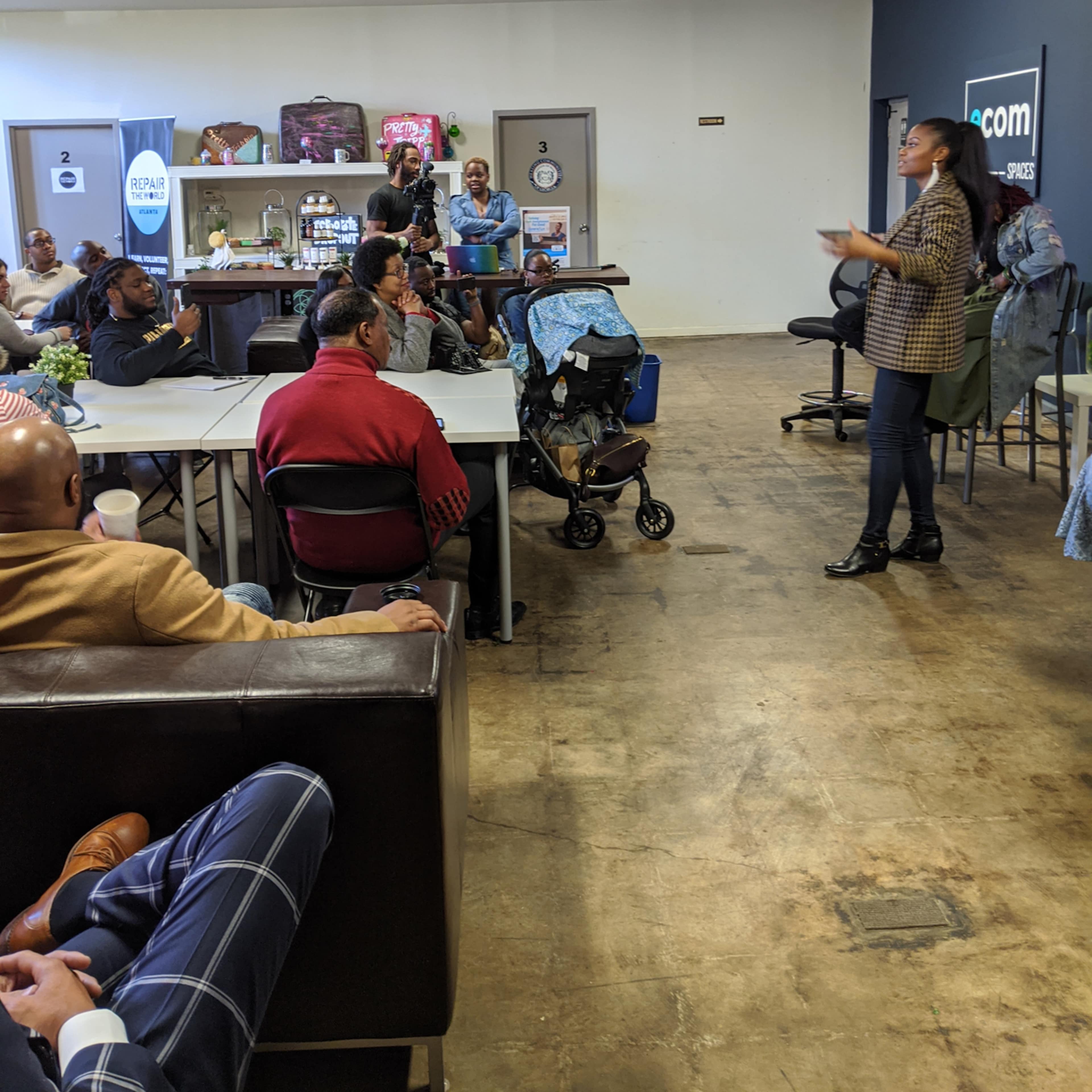 A woman speaks to an audience seated at tables in a communal space while others stand nearby.