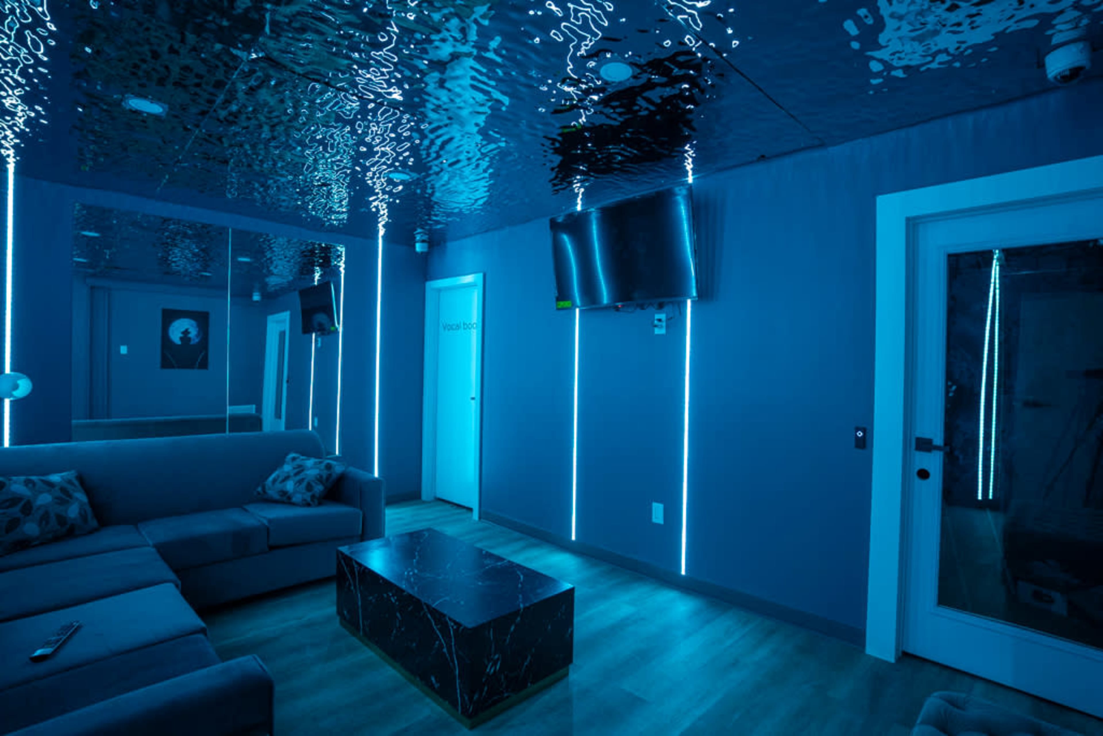A modern room with blue lighting, a sofa, a coffee table, a television mounted on the wall, and a door.
