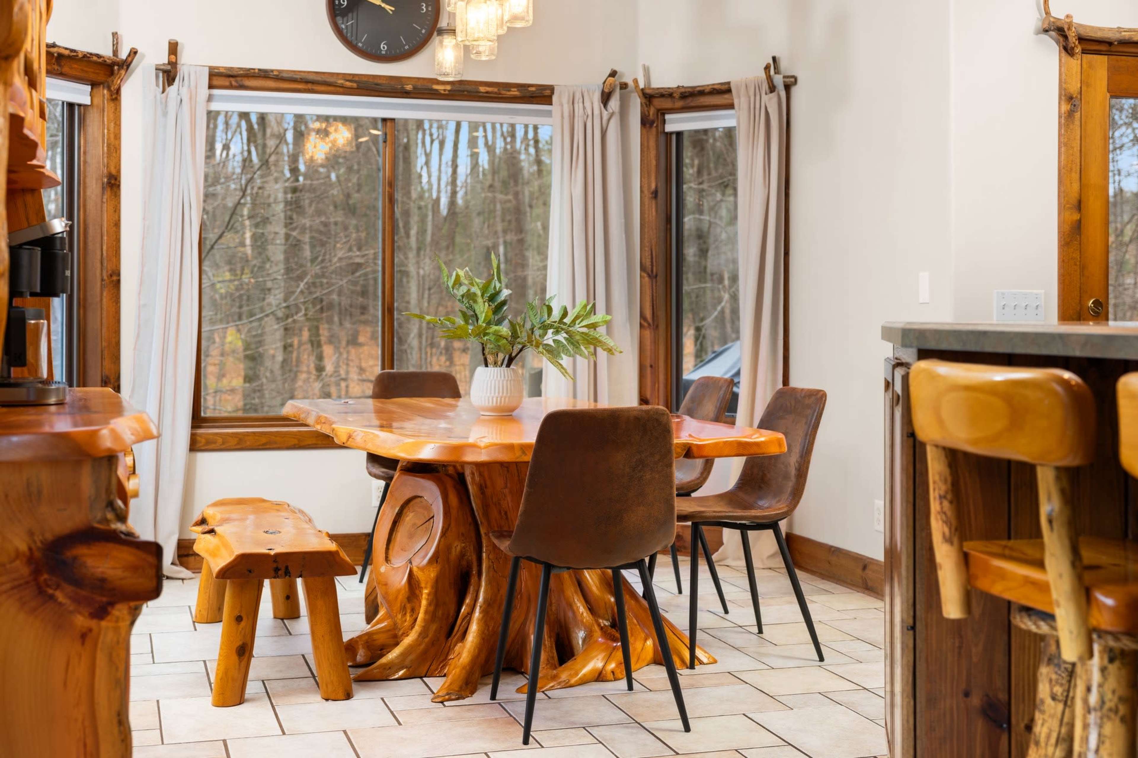 The image shows a dining area with a large wooden table surrounded by chairs, a bench, and large windows letting in natural light.