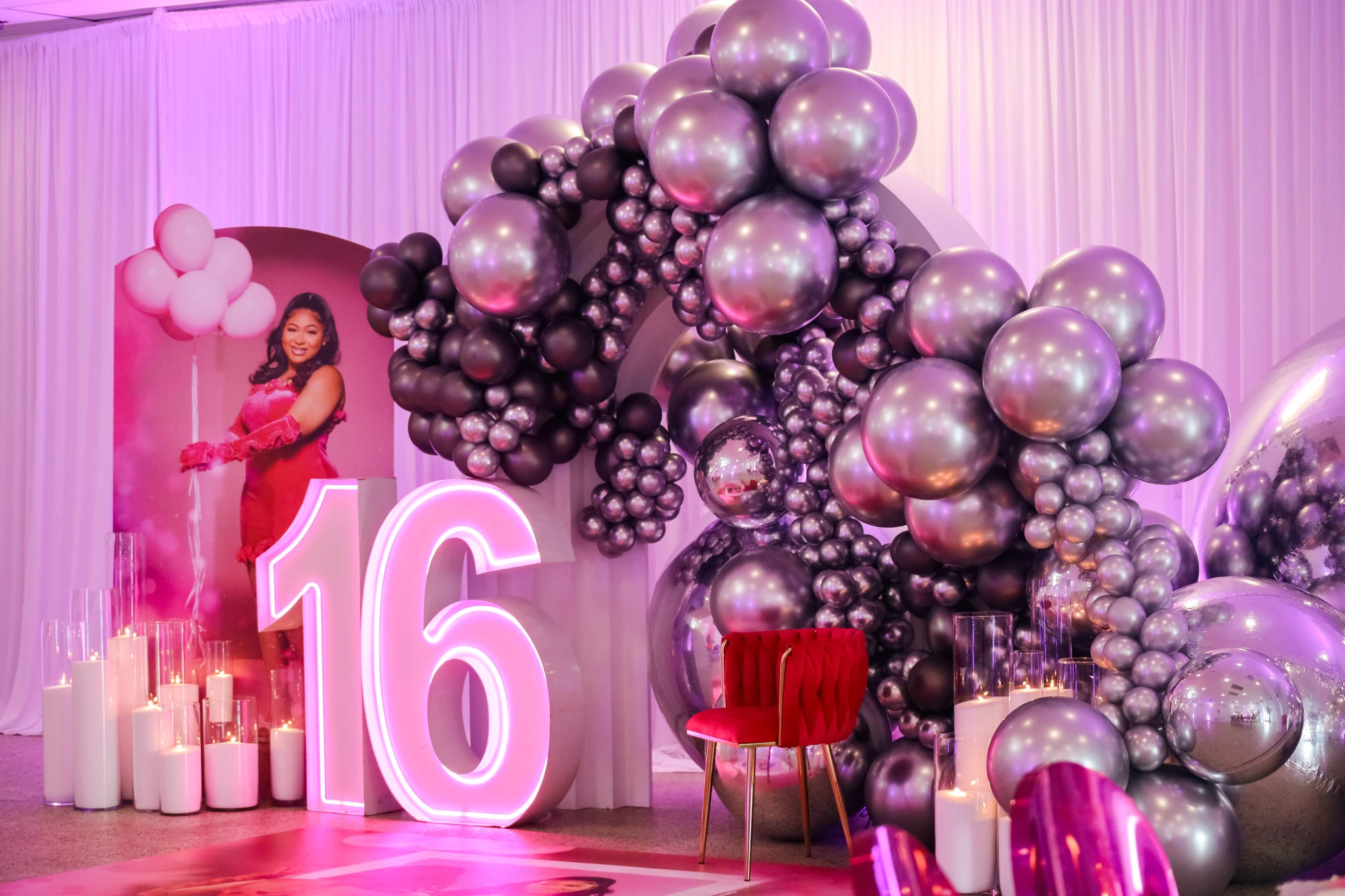 The image shows a festive event setup featuring large silver and black balloons, a prominent illuminated "16," and a red chair, with a person in a pink dress standing in the background.