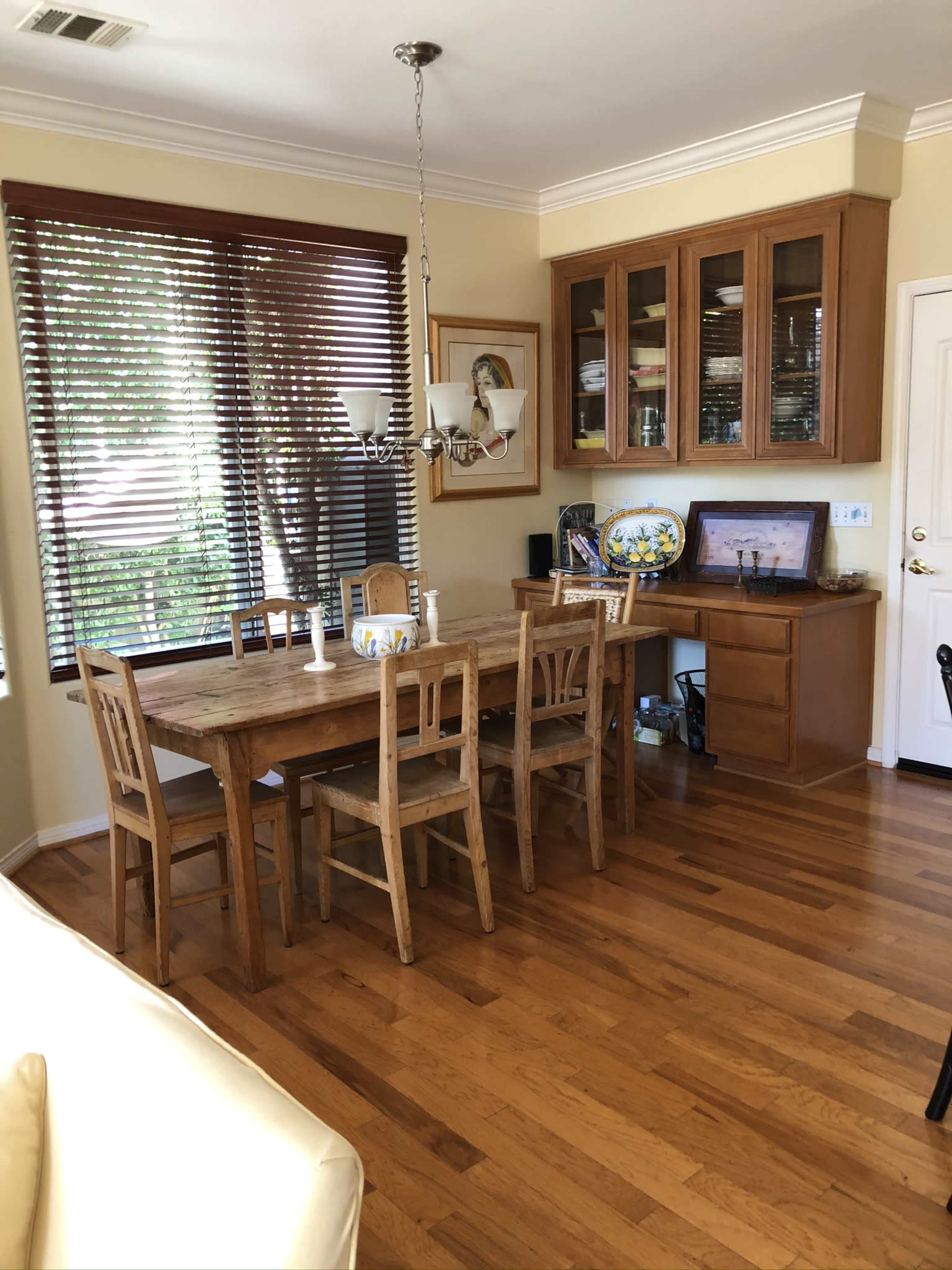 A dining area with a wooden table surrounded by six chairs, a china cabinet with glass doors, and a large window with blinds.