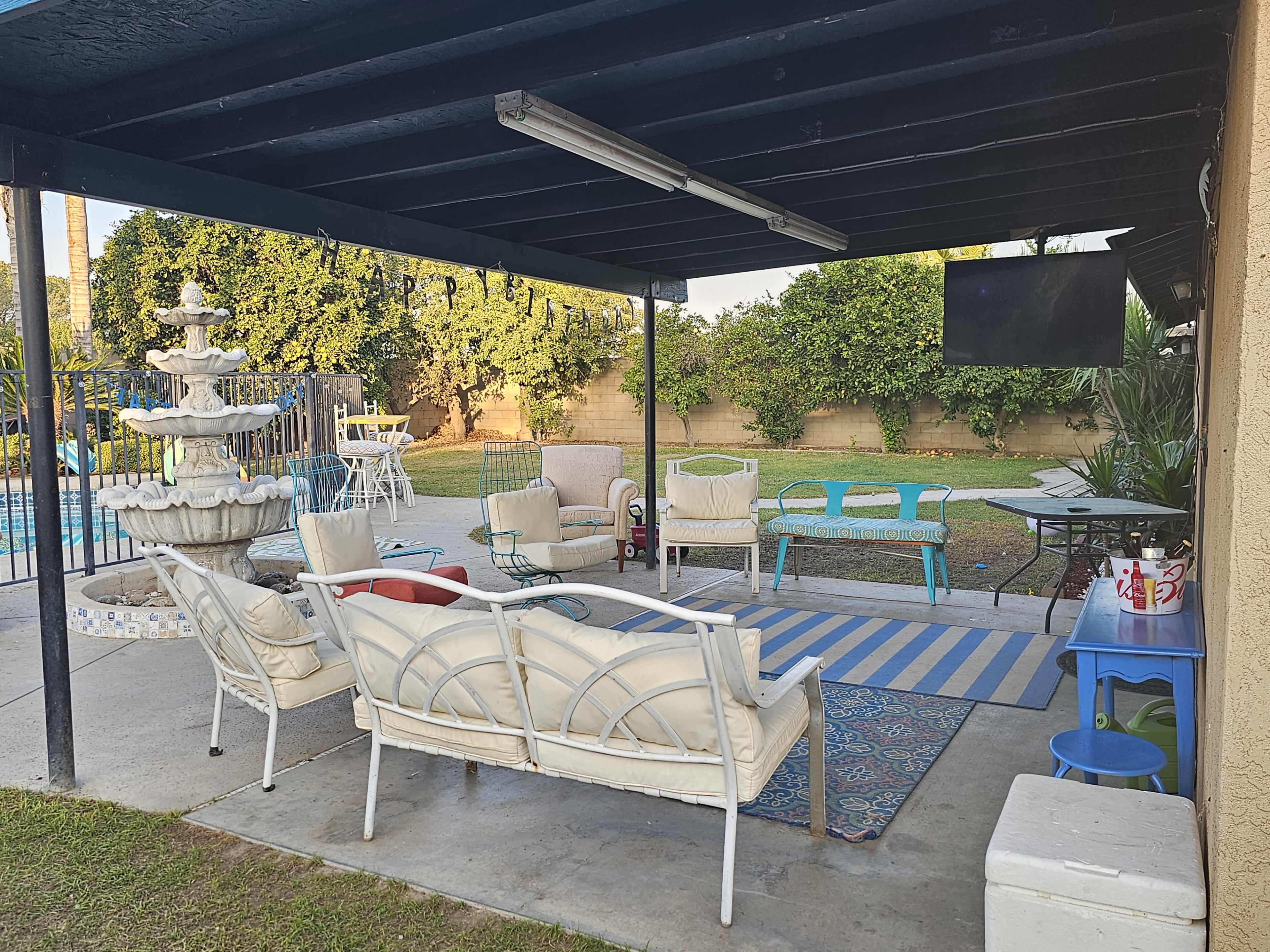 A covered outdoor seating area with white patio furniture, a fountain, a swimming pool in the background, and a television mounted on the wall.