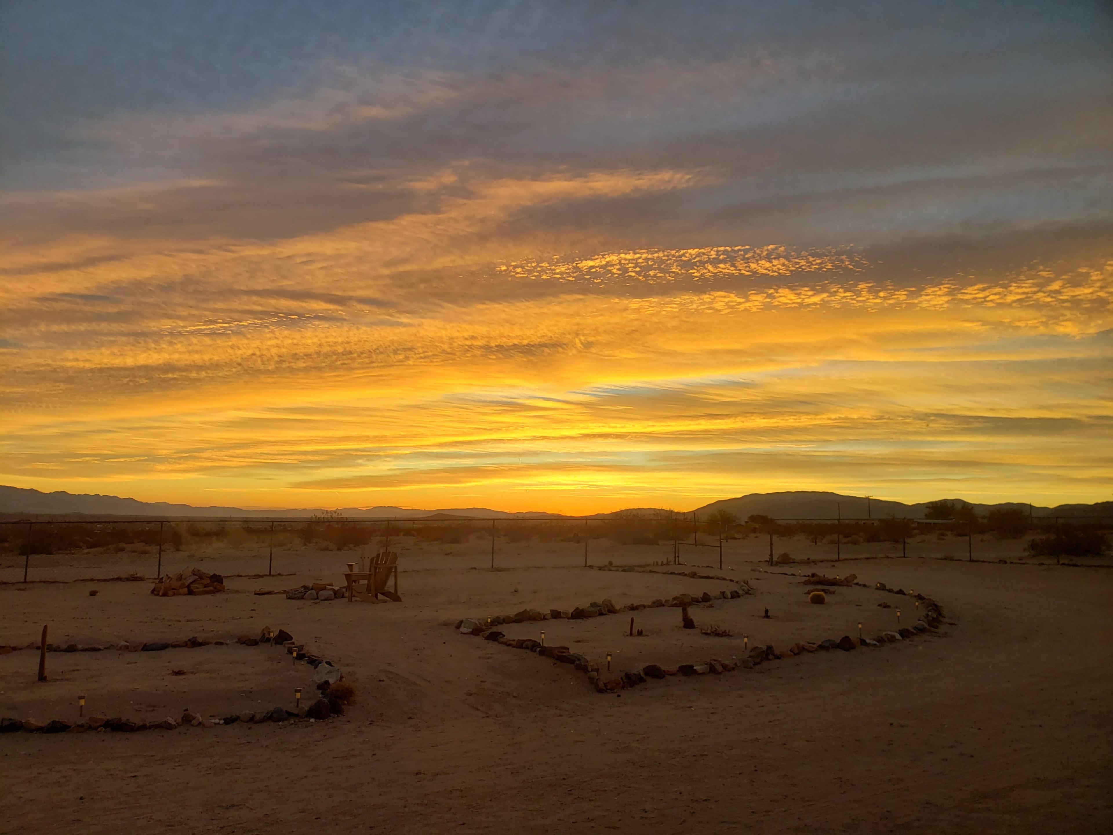 A desert landscape at sunset with a sky filled with yellow and orange hues, and a circular arrangement of stones on the ground.