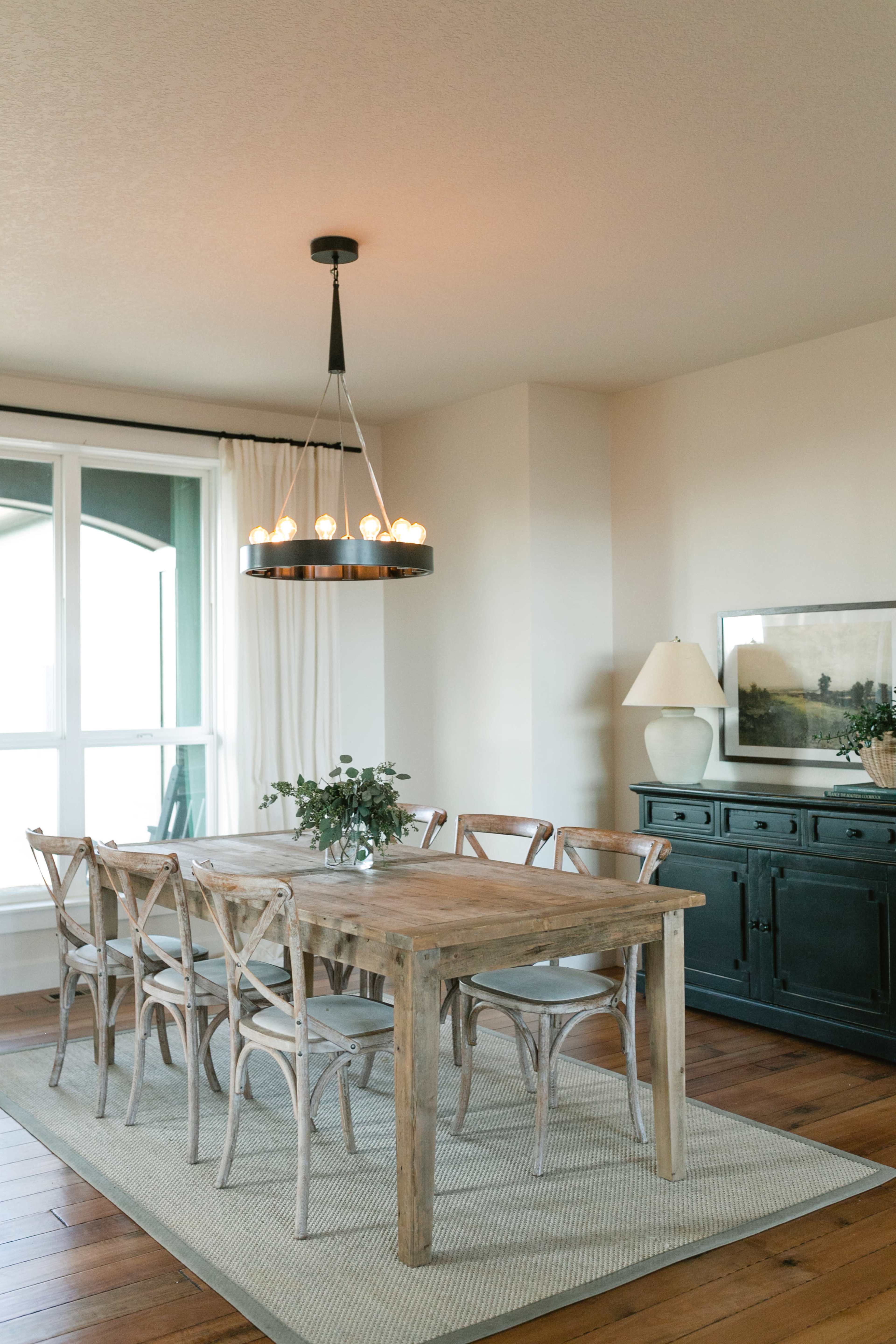 A spacious dining room features a large wooden table surrounded by six chairs, a modern chandelier above, and a sideboard against the wall.