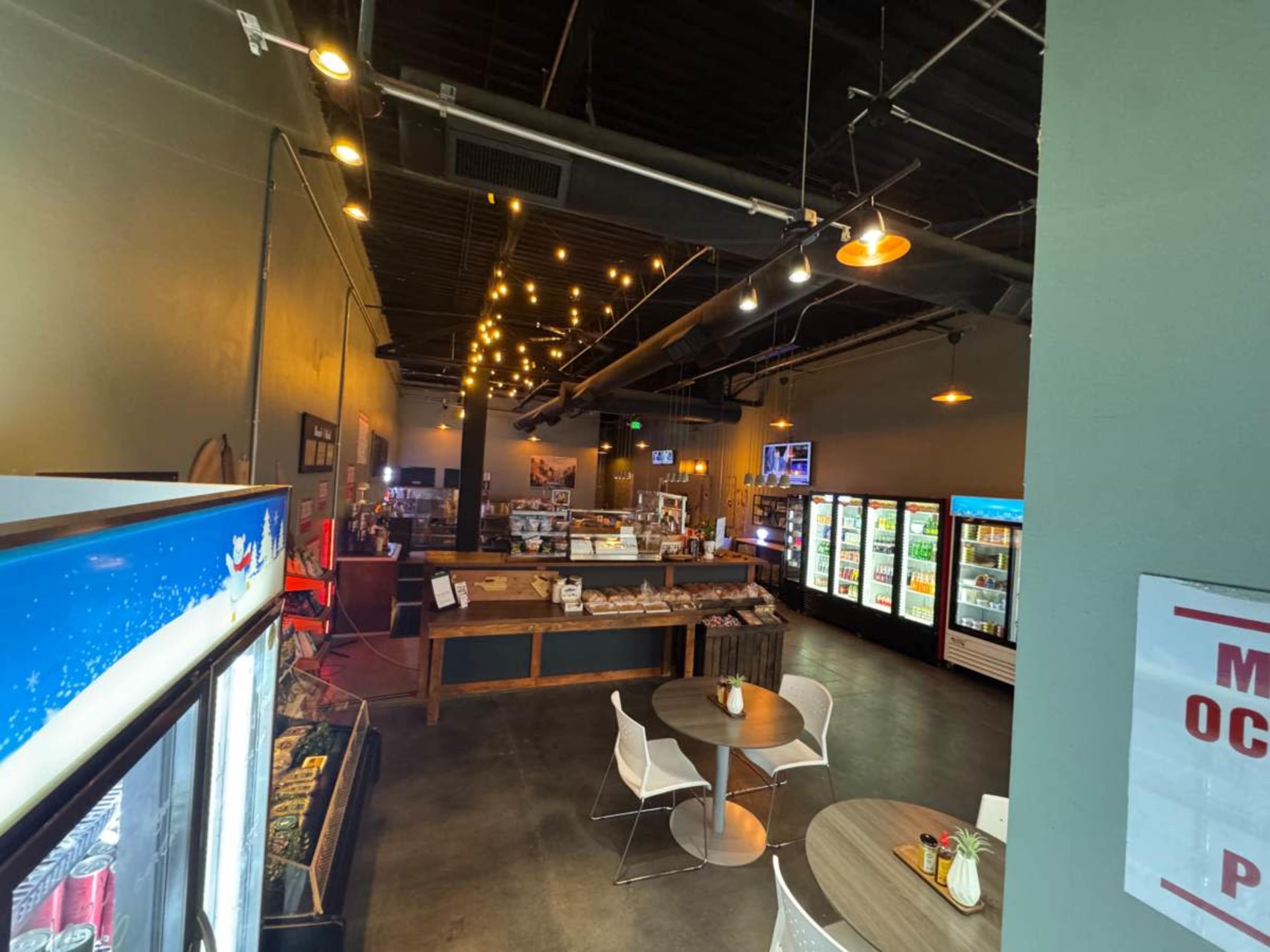 The image shows a spacious café or store interior with tables, a service counter, and several refrigerators stocked with beverages.