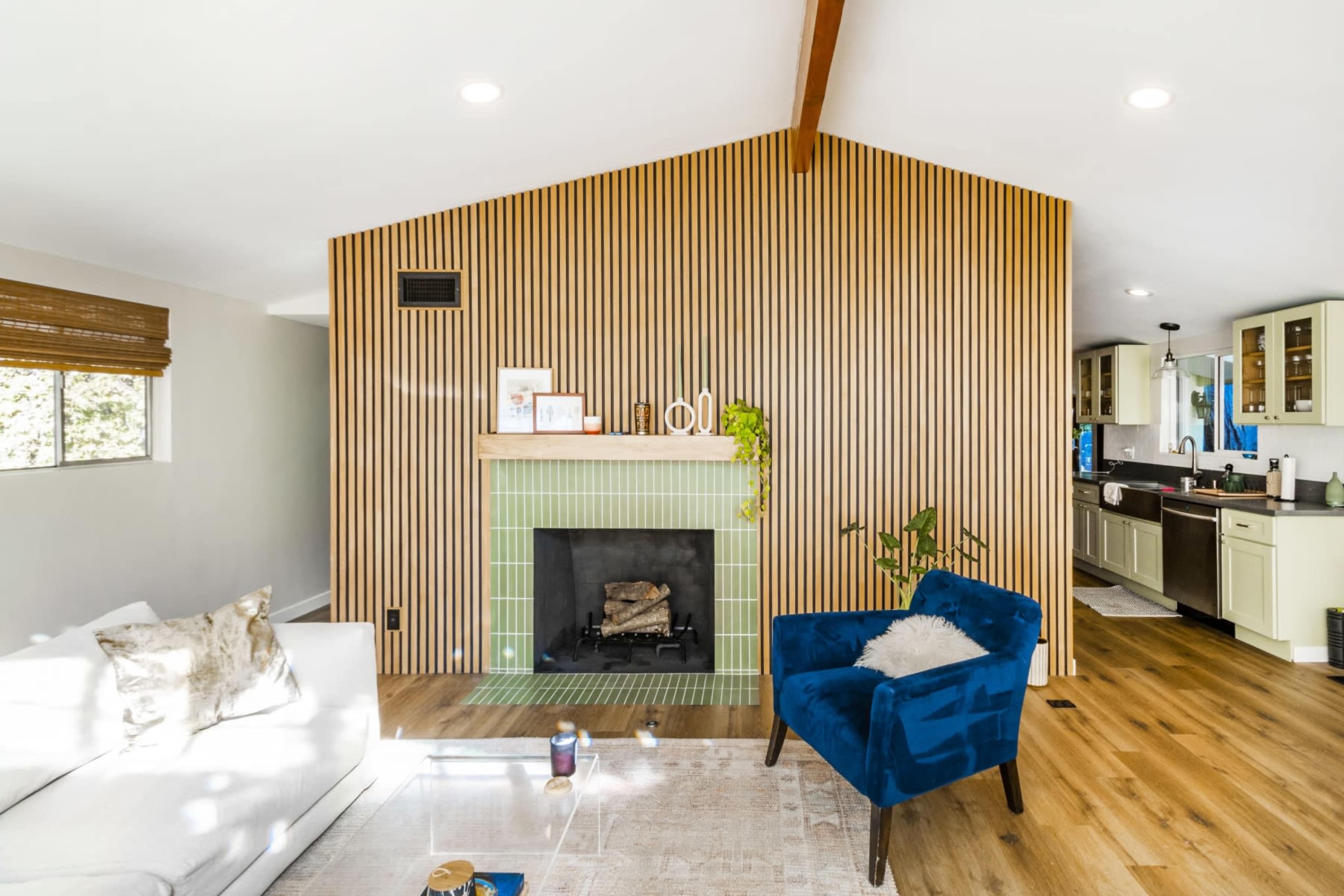 A modern living room featuring a wooden slat wall behind a green tiled fireplace, with a white sofa and a blue armchair in the foreground.