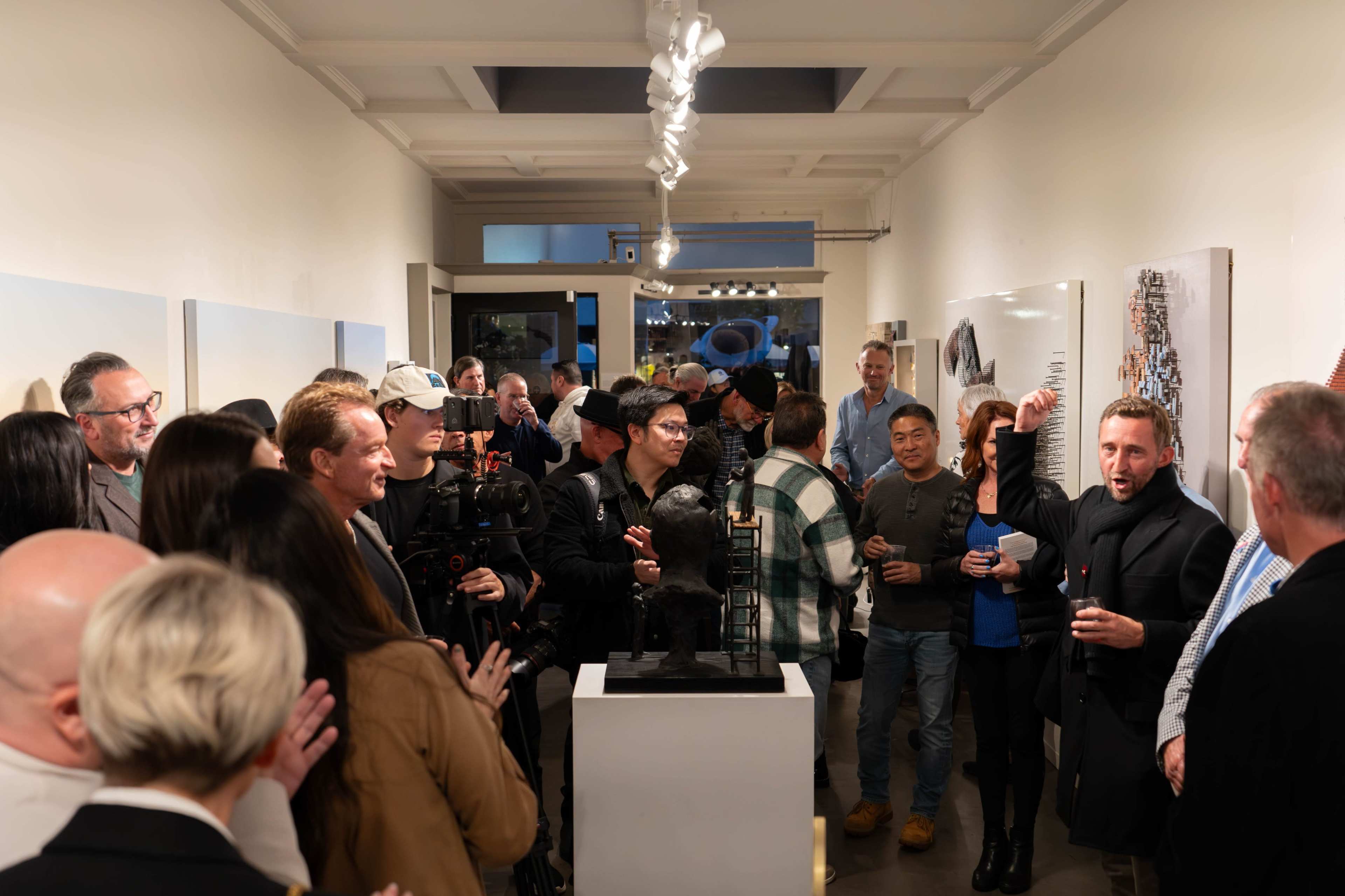A large crowd of people is gathered in an art gallery, engaging in discussions while looking at various artworks on the walls.