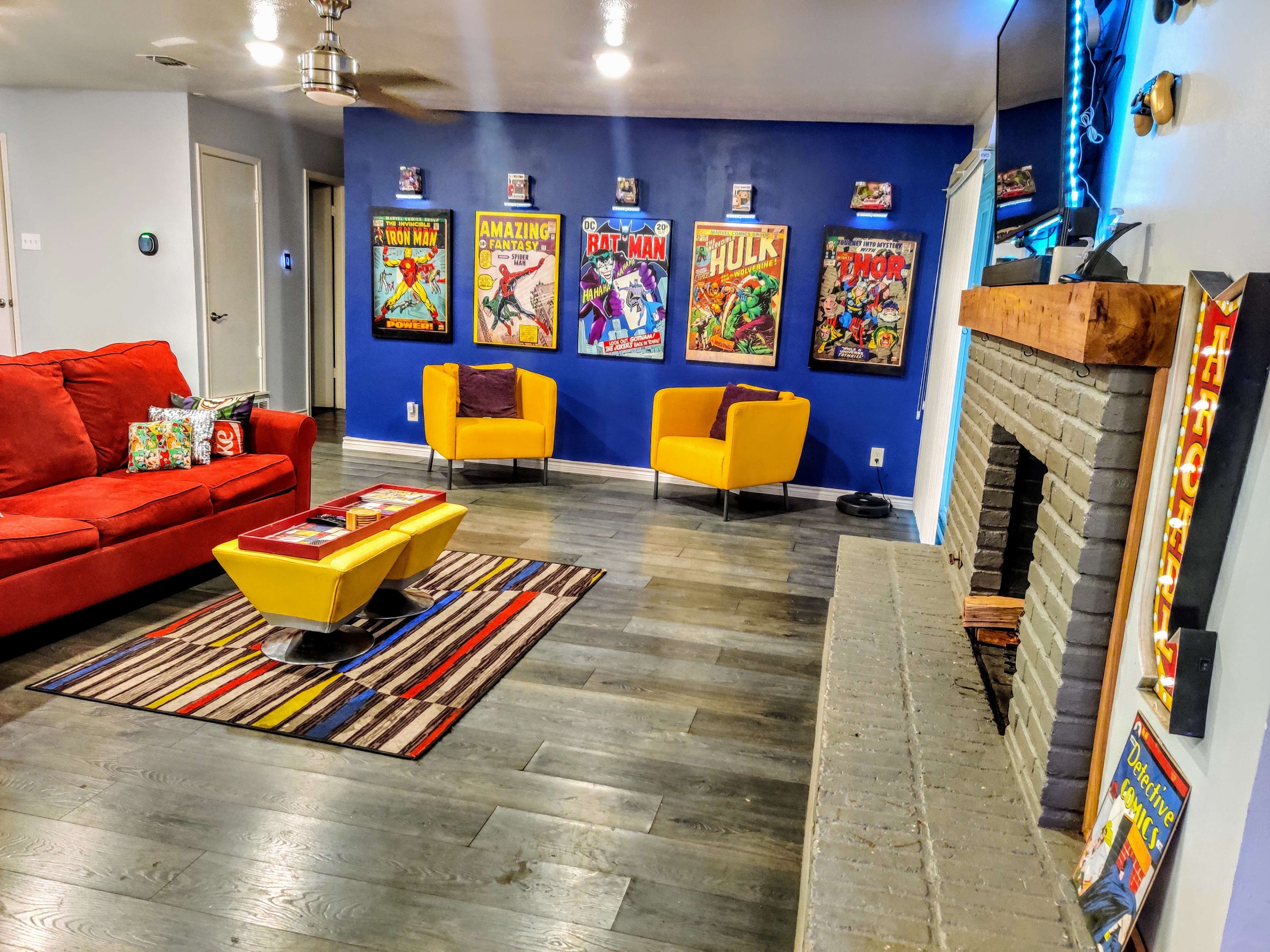 The living room features a red sofa, yellow chairs, a patterned rug, and a wall adorned with framed comic book posters.