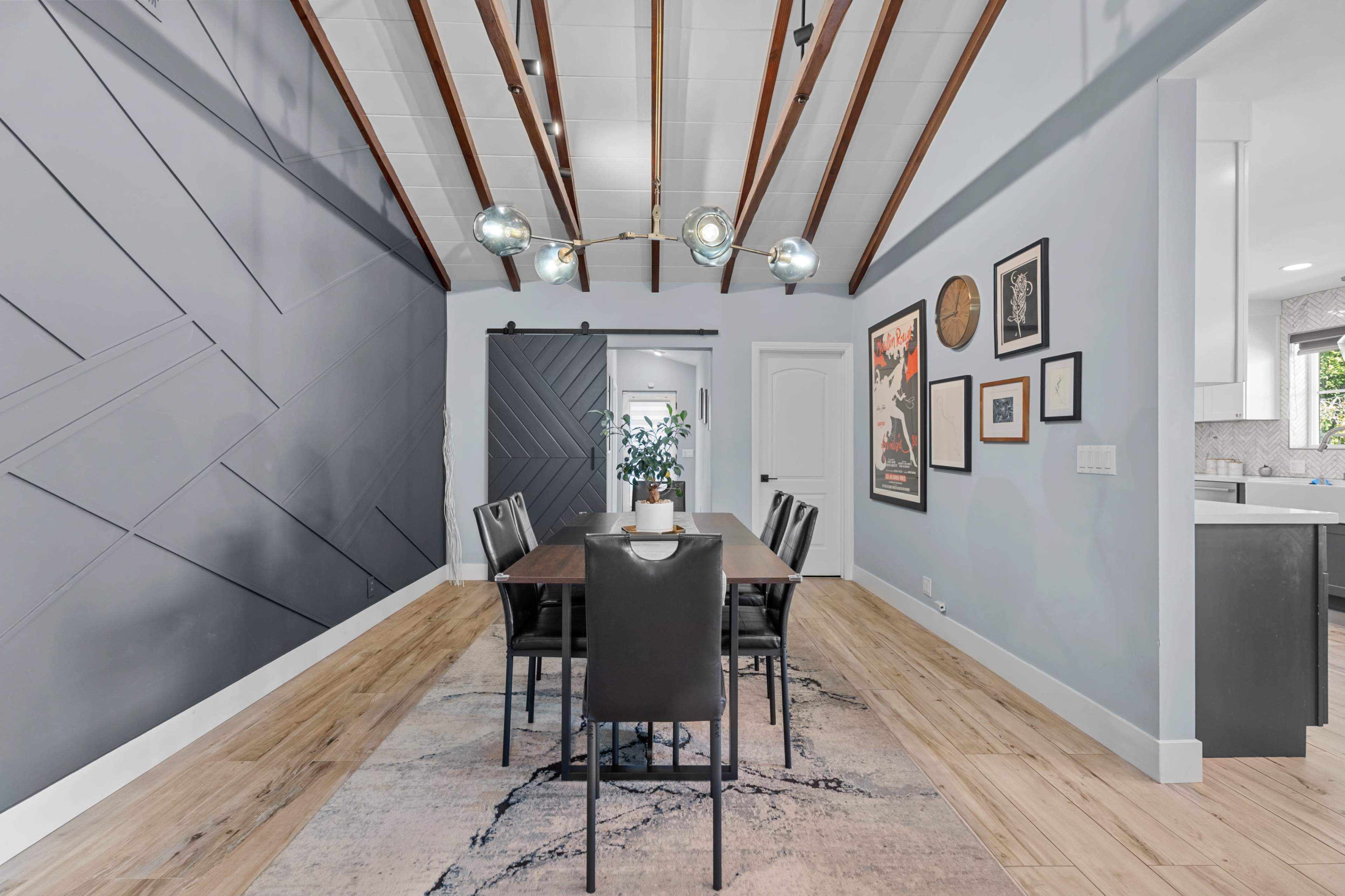 The image depicts a dining area with a long table surrounded by chairs, featuring a textured wall and exposed wooden beams overhead.