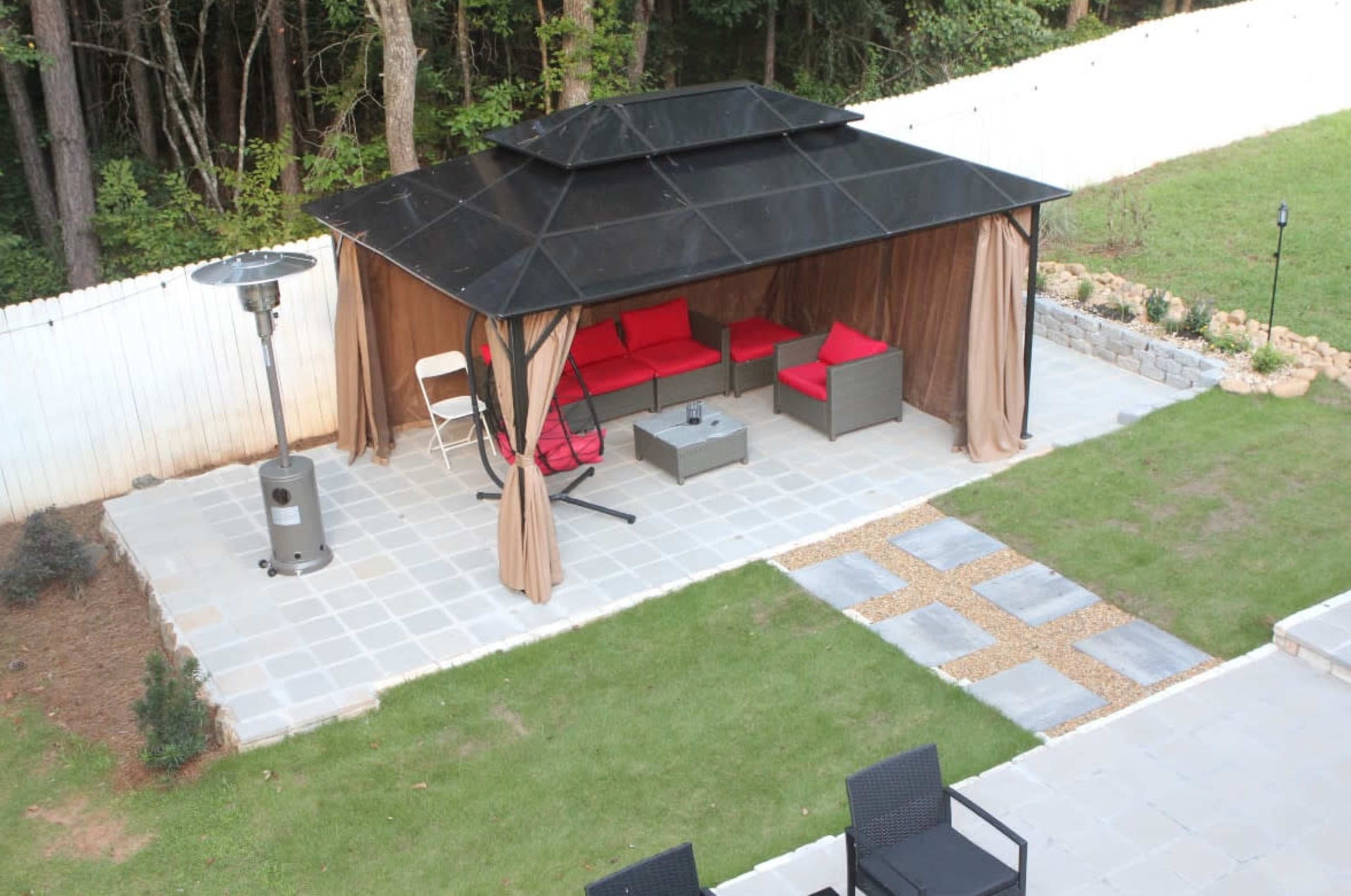 The image shows a gazebo with red seating, a table, and a patio heater set on a paved area in a backyard surrounded by trees.