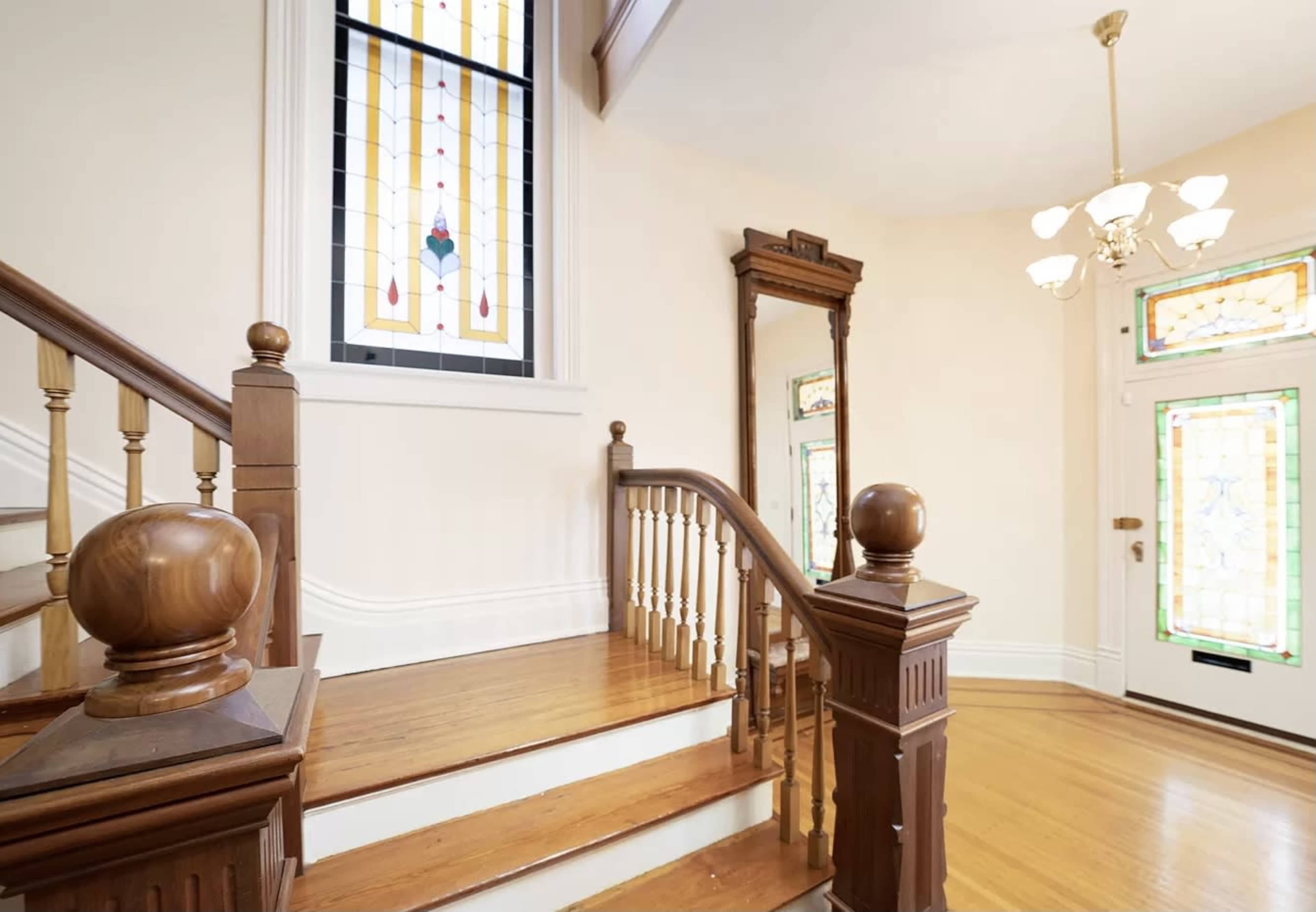 A well-lit interior with a wooden staircase leading to an upper floor, framed by stained glass windows and a chandelier hanging from the ceiling.