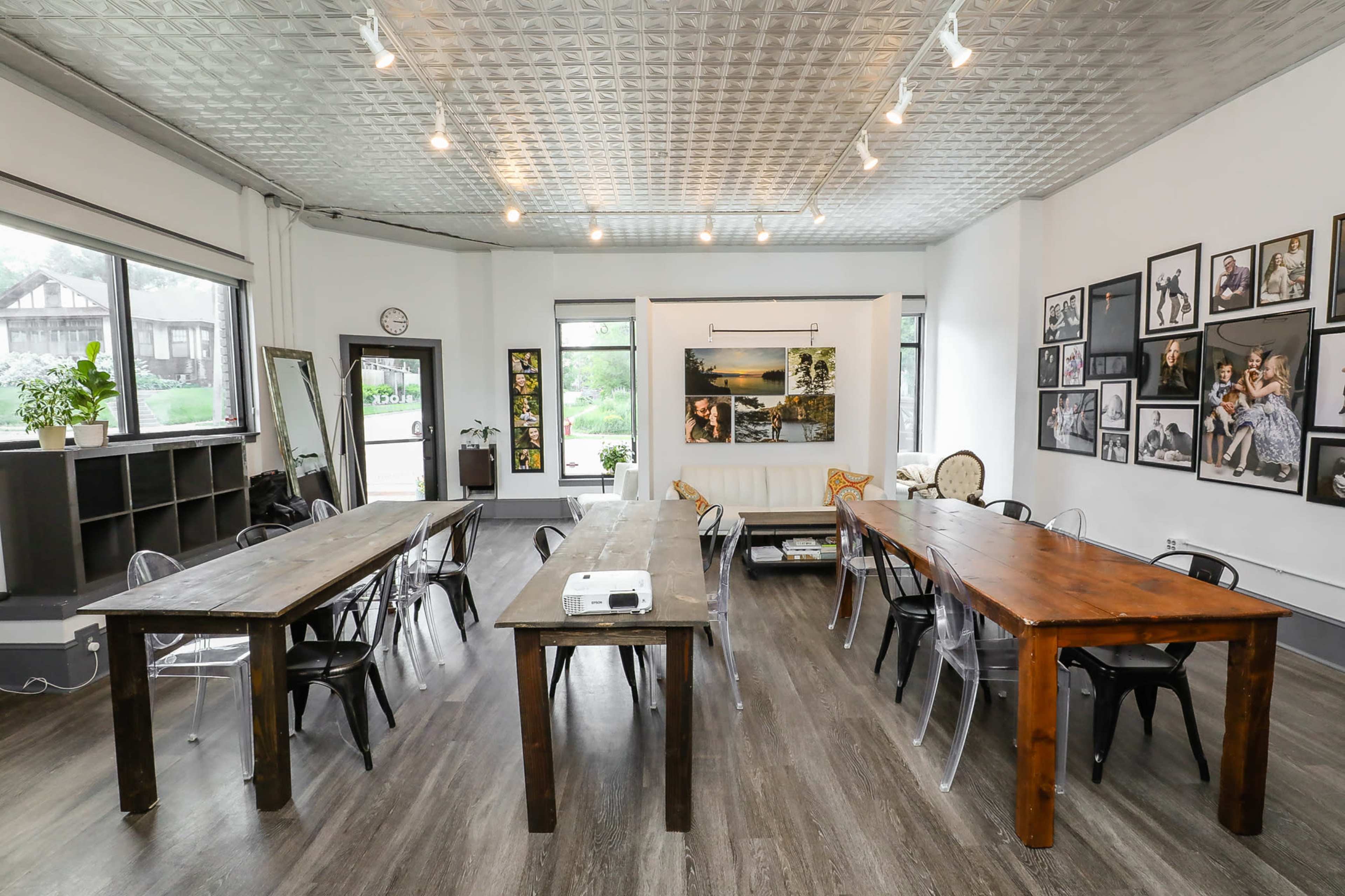 The image shows a spacious room with two wooden tables, clear chairs, and various framed photographs on the walls.