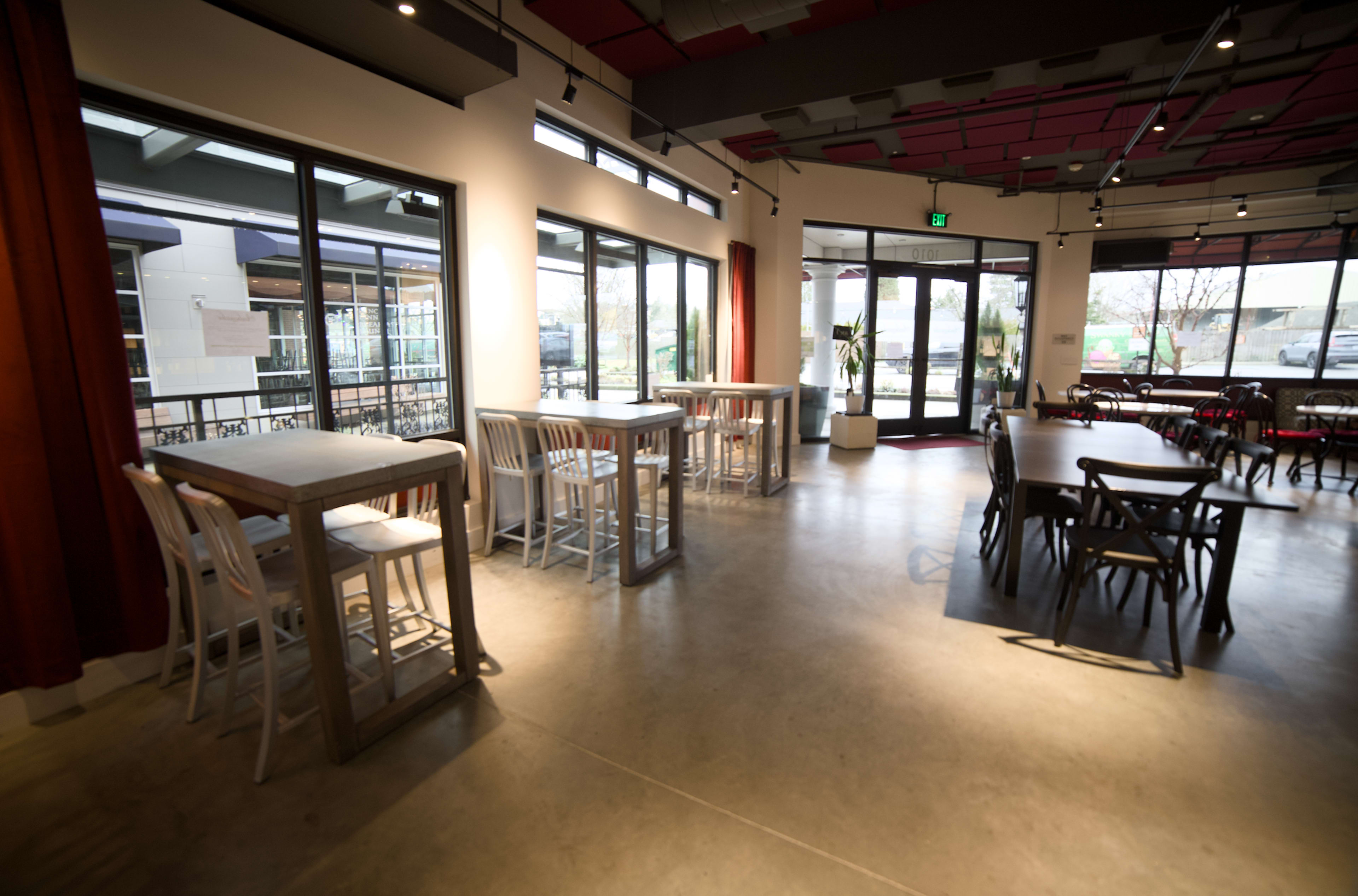 The image shows a modern café interior with a mix of high tables and standard dining tables, large windows allowing natural light, and minimalistic décor.