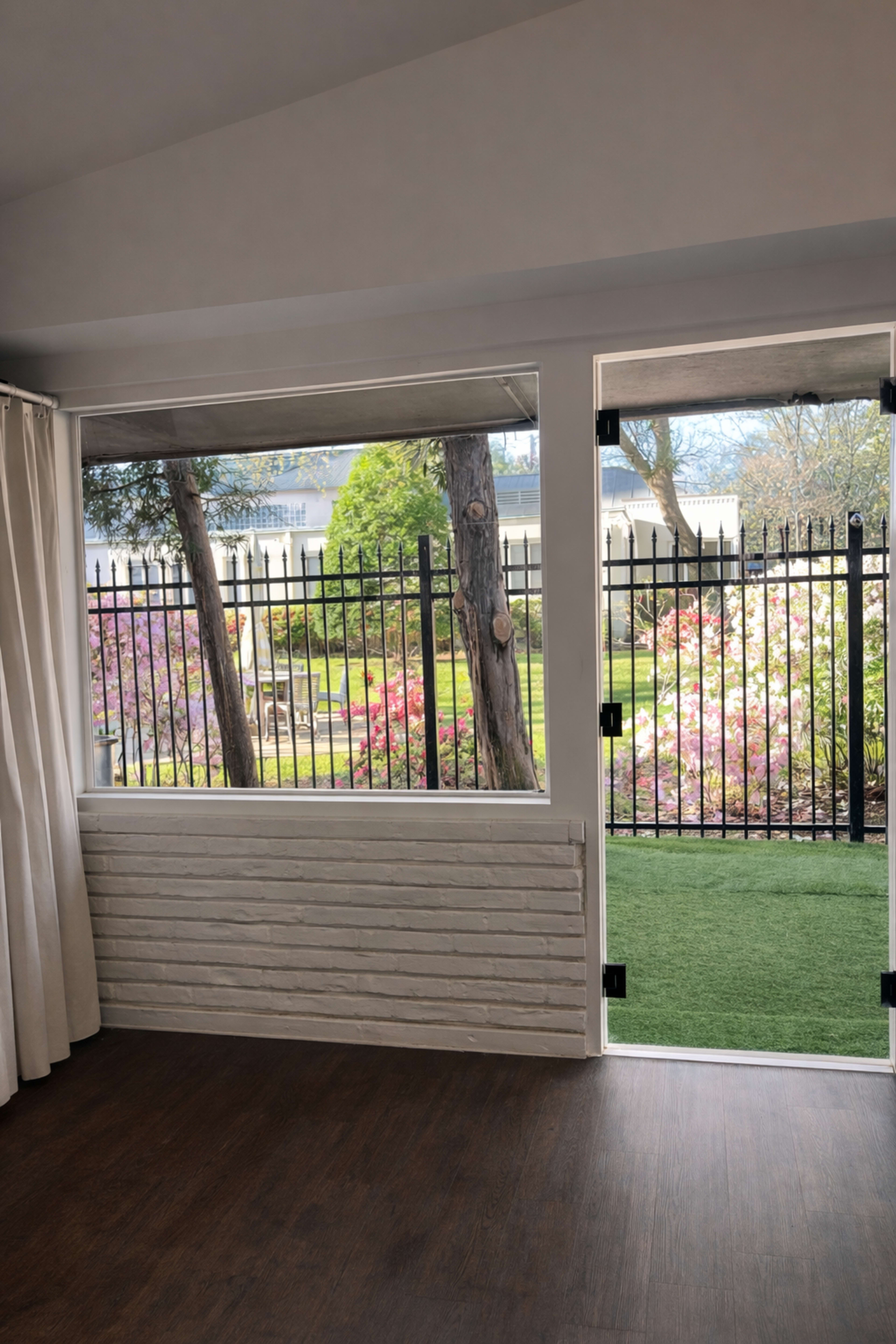 The image shows an interior room with a window and door leading to a fenced outdoor area featuring greenery and blooming flowers.