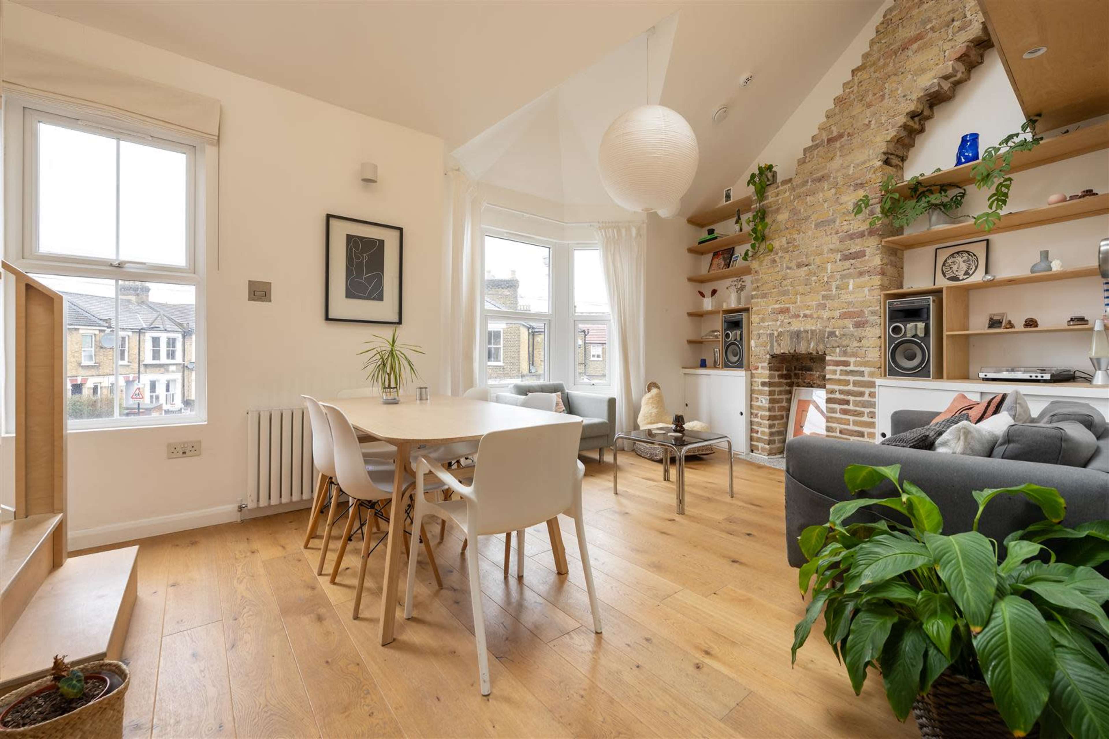 A modern living space features a dining area with a wooden table and white chairs, complemented by a cozy lounge with a gray sofa, exposed brick wall, and large windows.