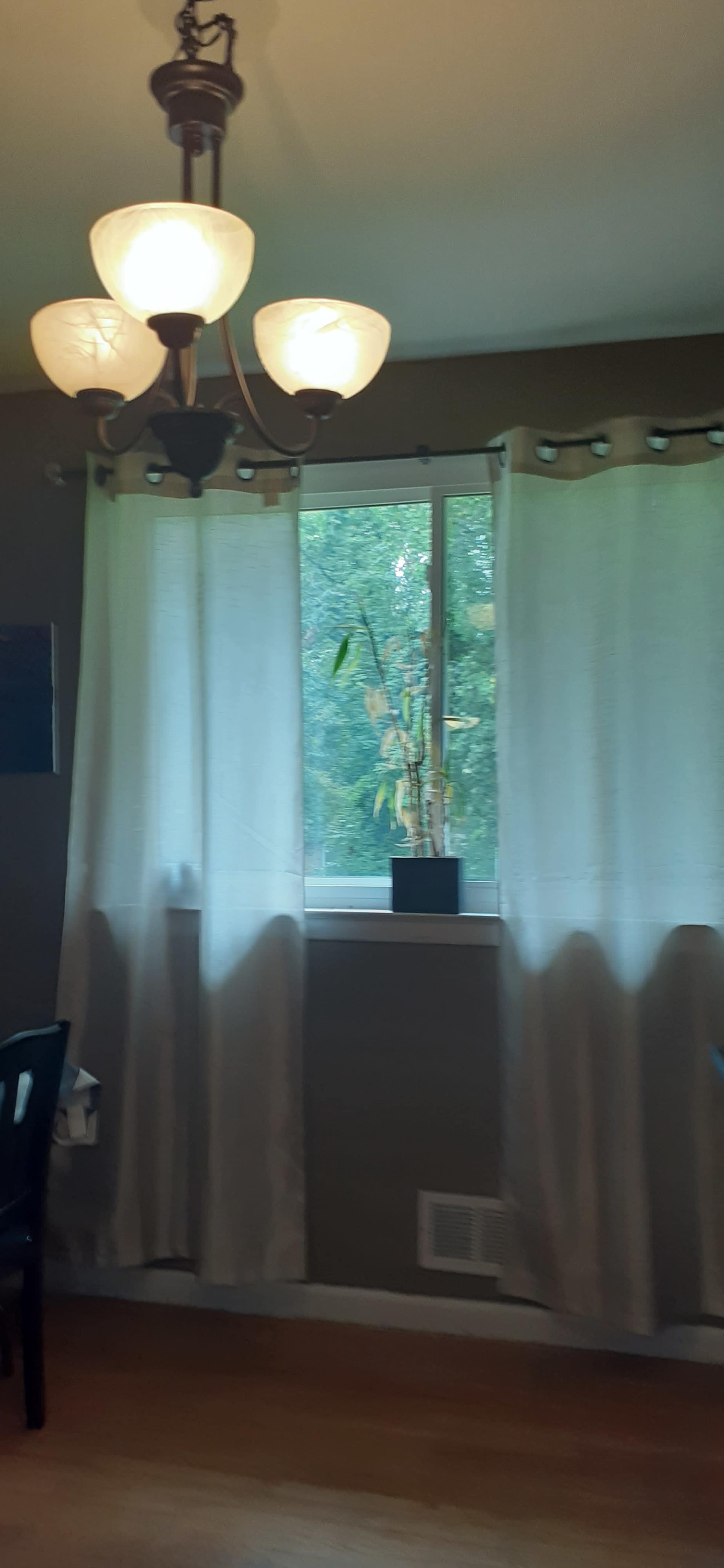 A window with sheer curtains and a small potted plant is set in a room featuring a chandelier and a dark table.