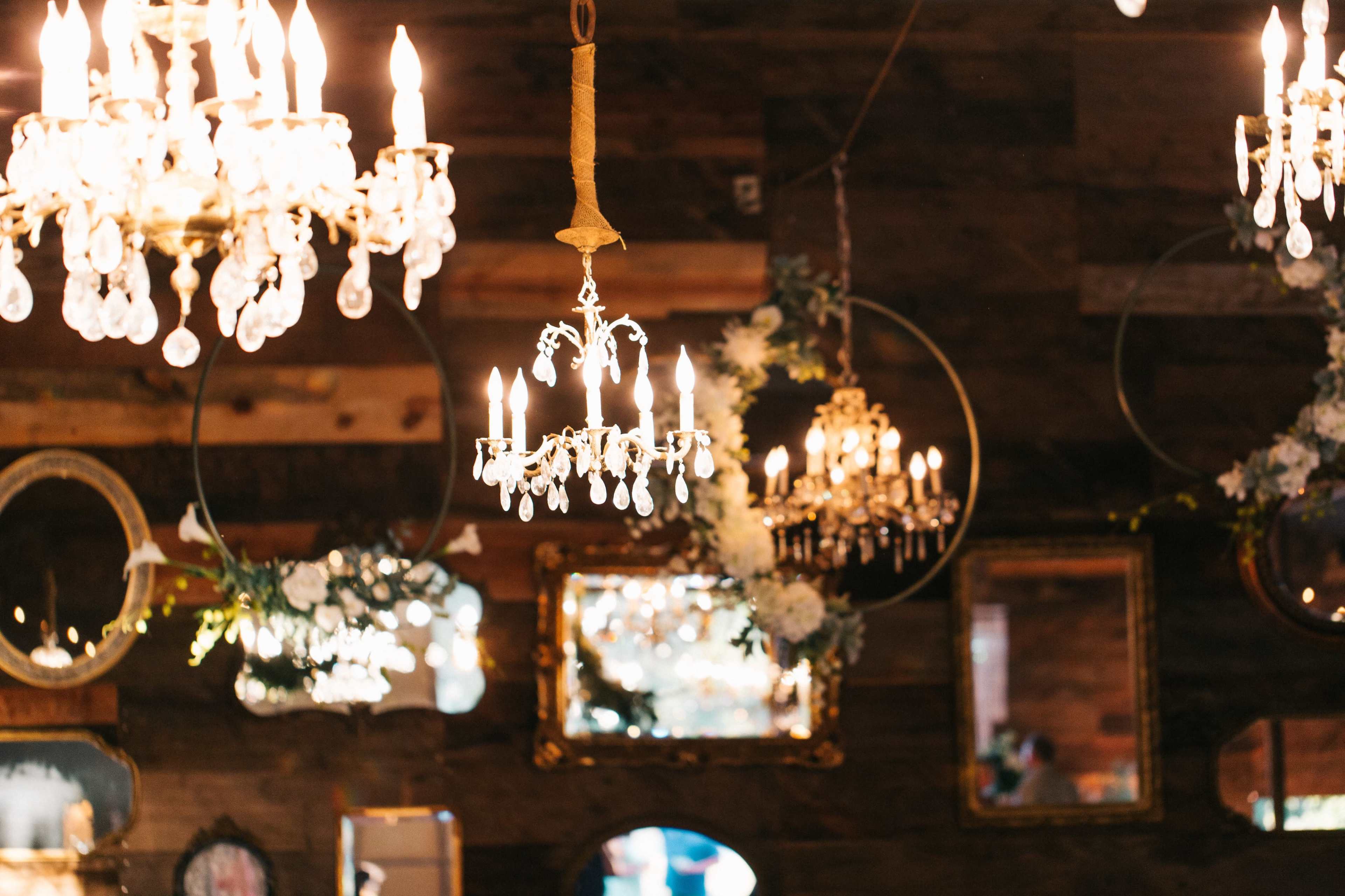 The image shows an array of chandeliers hanging from a wooden ceiling, surrounded by decorated mirrors and floral accents.