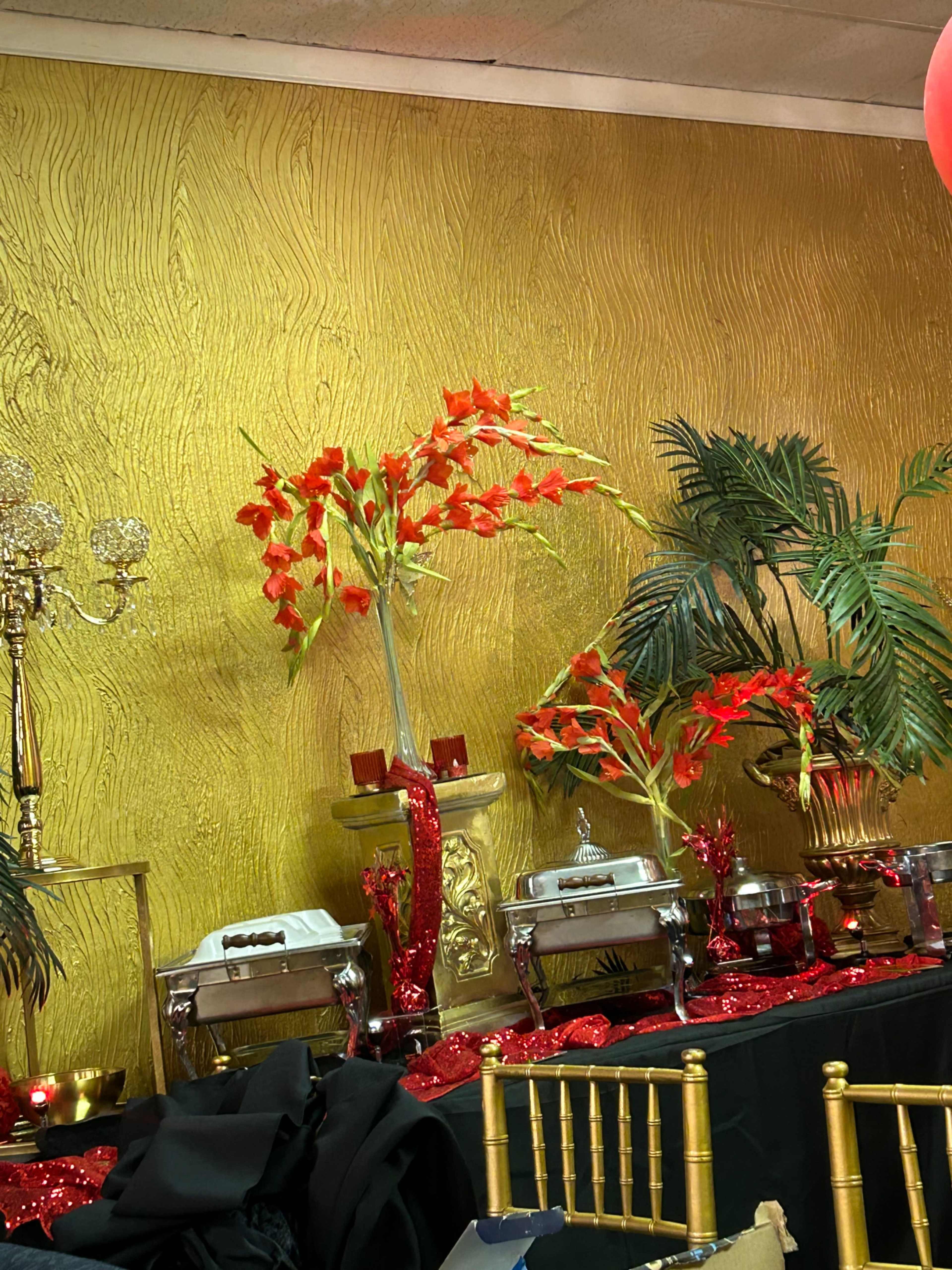 A vibrant gold wall serves as a backdrop to a display of decorative red flowers and elegant serving dishes set on a table adorned with red sequins.