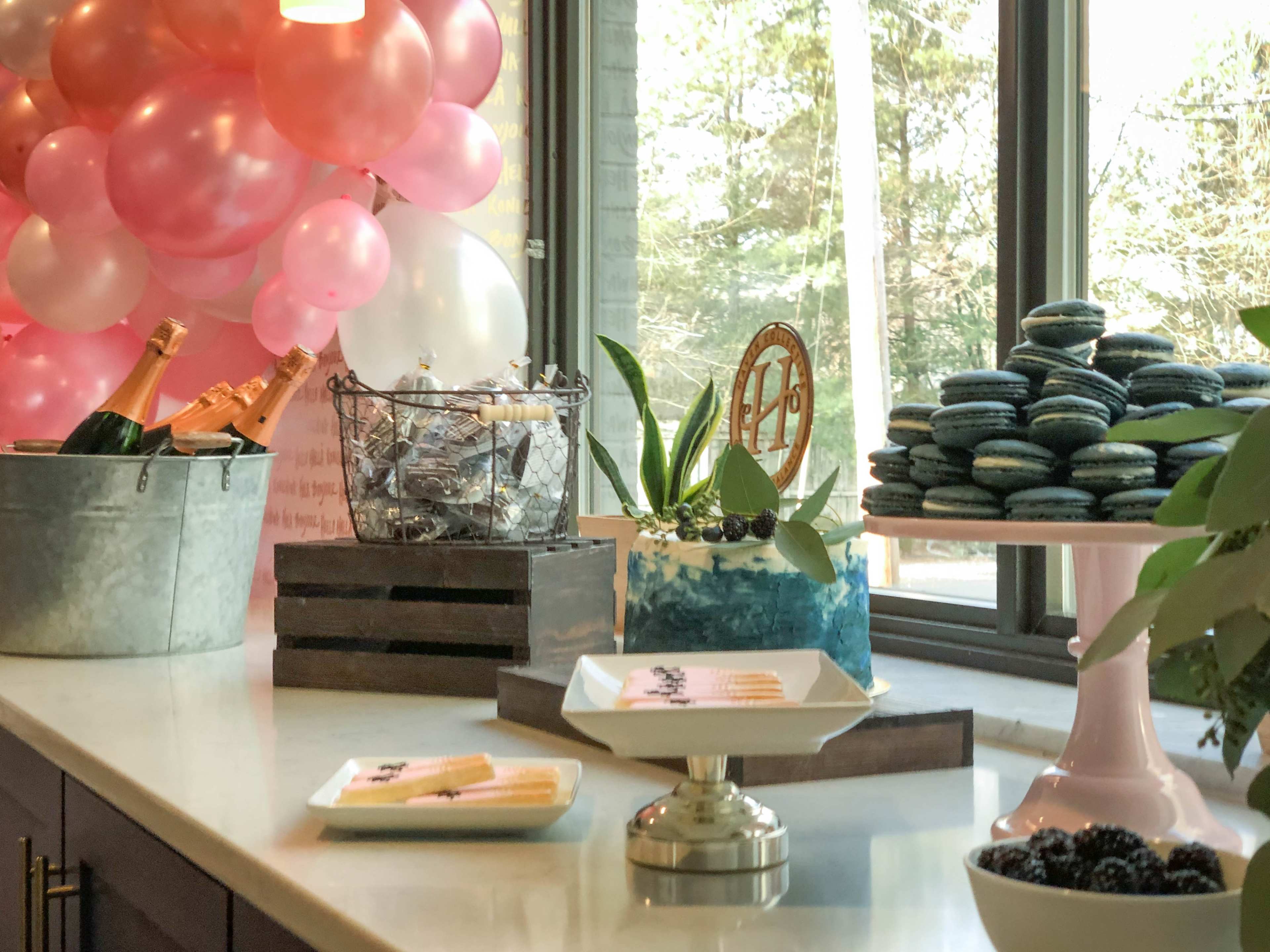 A table is set with a decorated cake, macarons, champagne bottles, and a balloon backdrop in a brightly lit room.